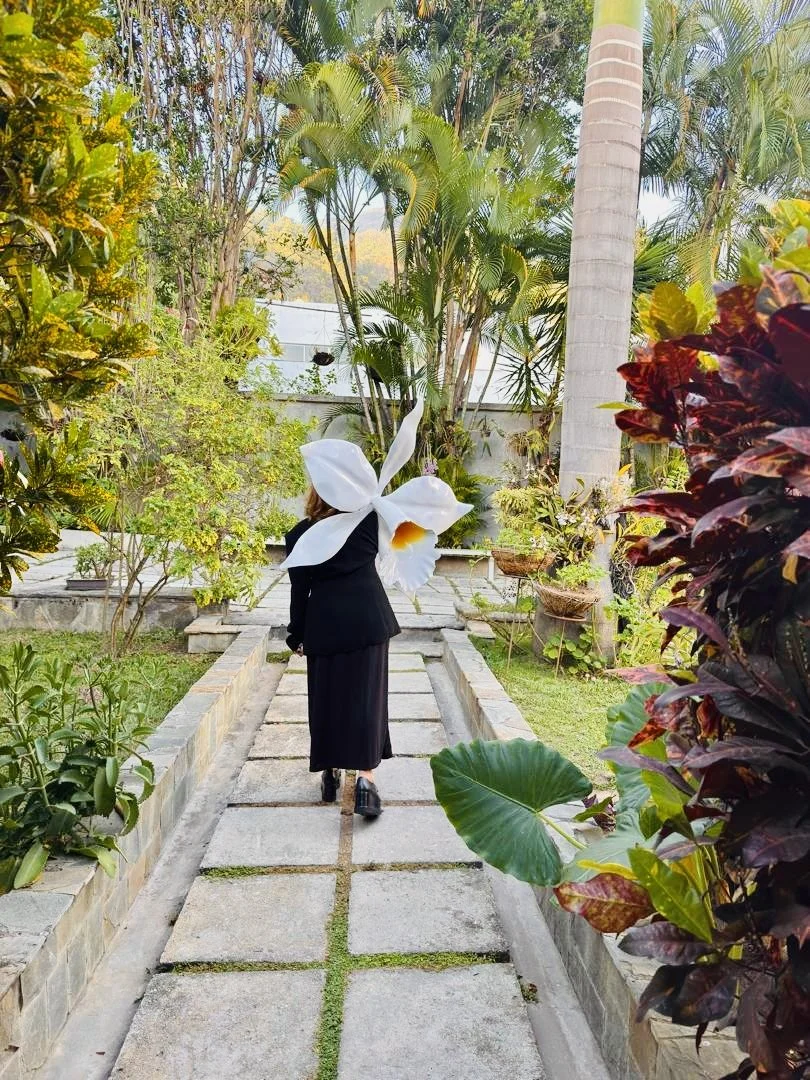 Person in black clothing walking on a stone pathway in a lush garden with tropical plants and trees, carrying a large white flower-like decoration on their back.
