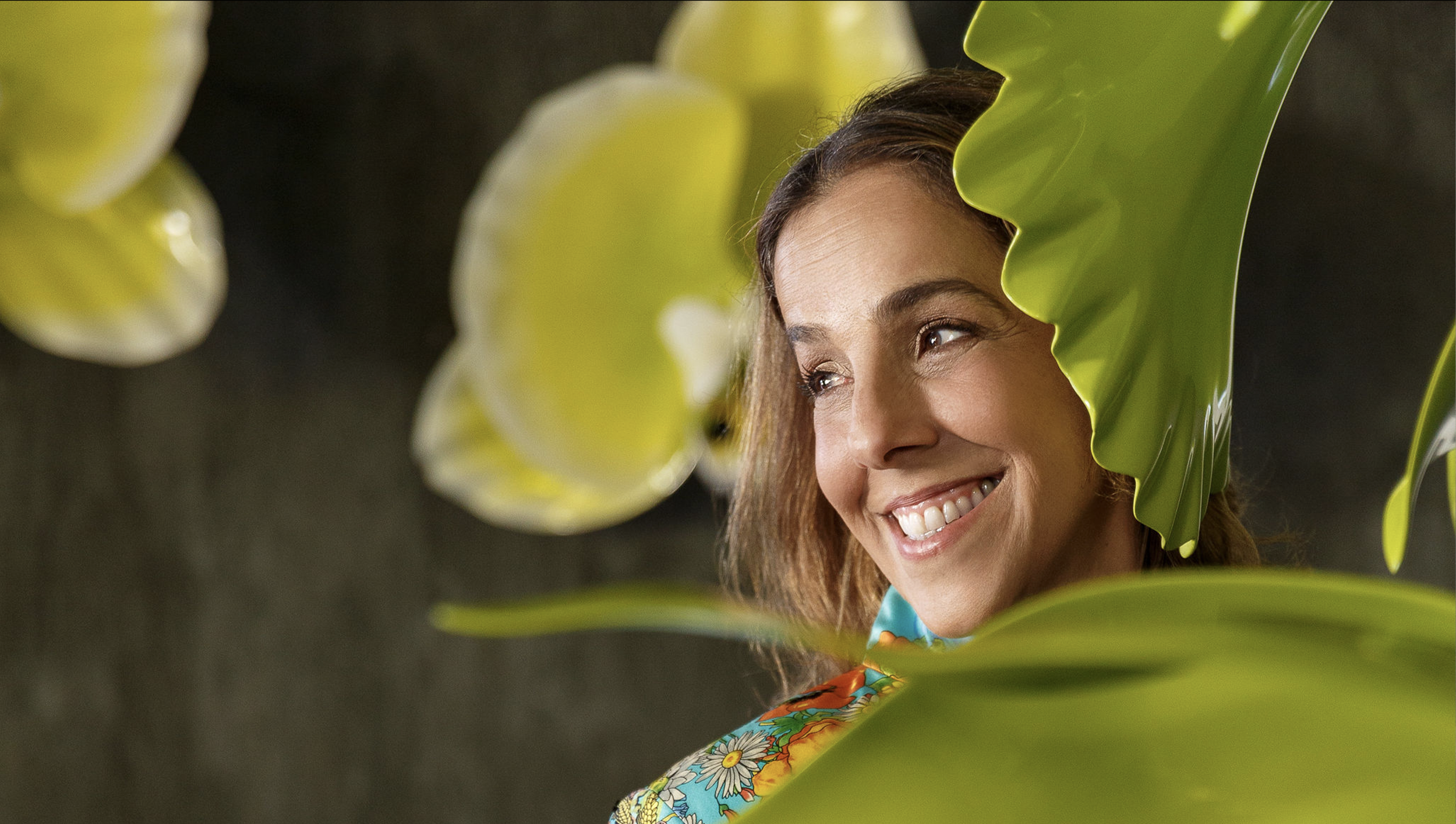 A woman with long hair smiling behind large green leaves.