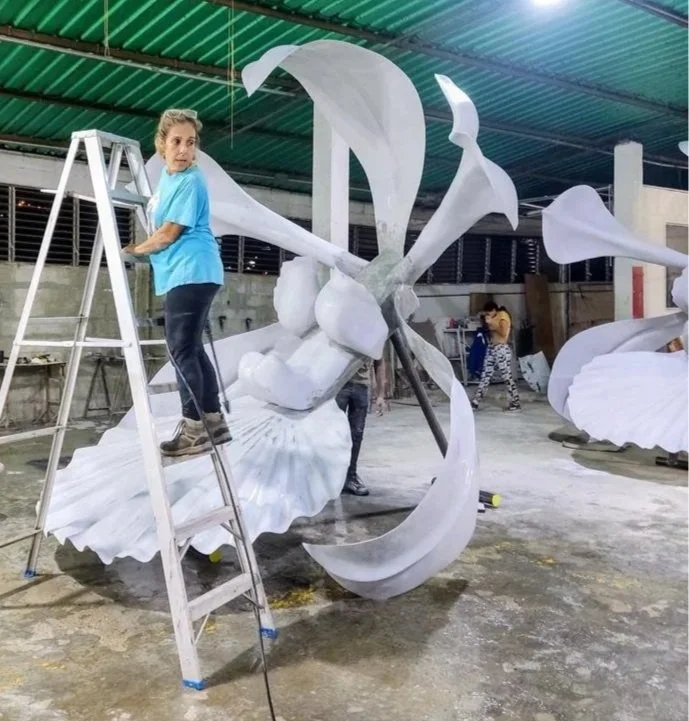 A woman on a ladder is working on a large white flower sculpture, with other people and sculptures in the background inside a workshop or warehouse.