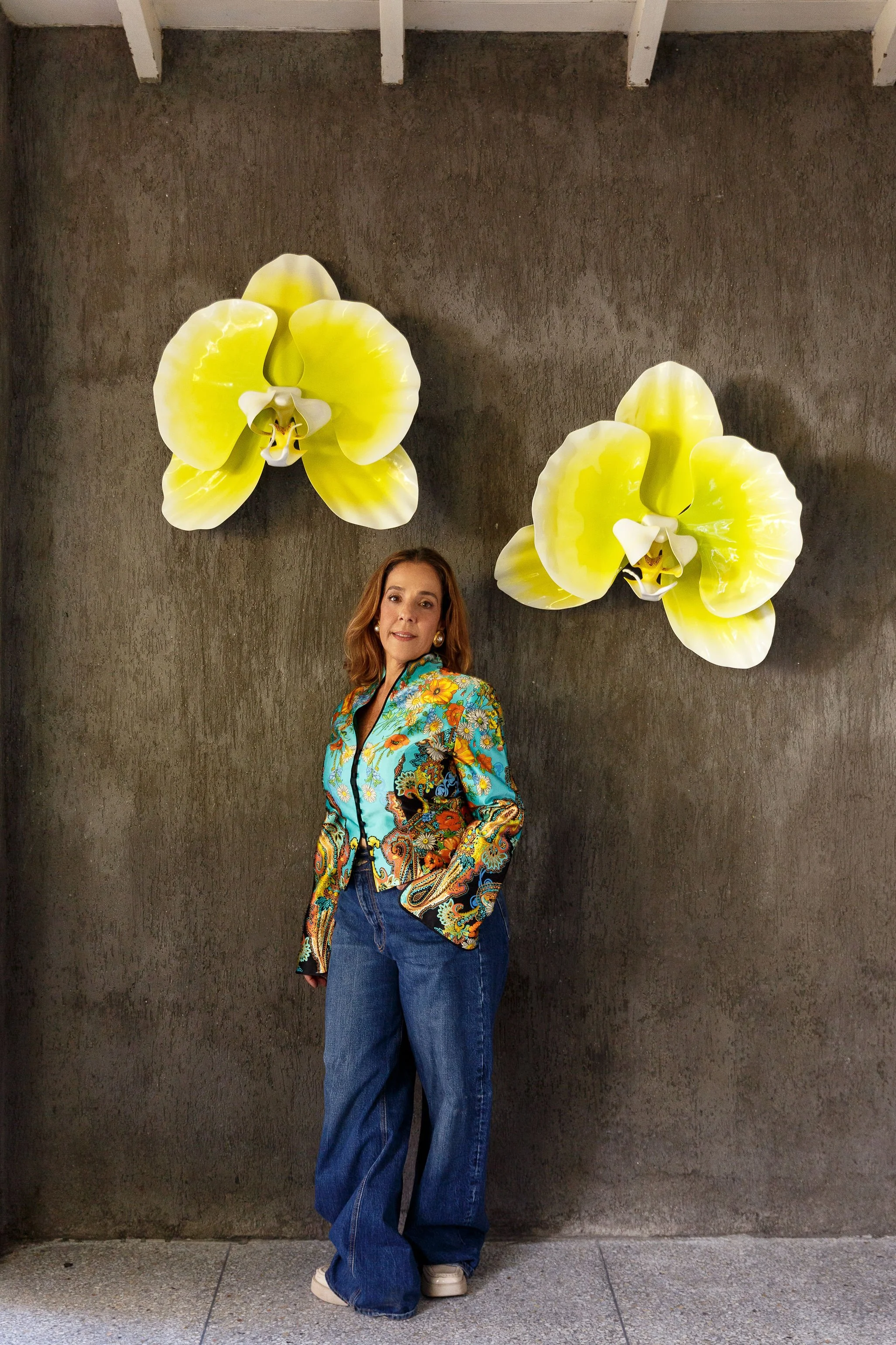 A woman in a colorful, patterned jacket and high-waisted jeans posing in front of a dark gray textured wall with two large yellow orchid flower wall decorations.