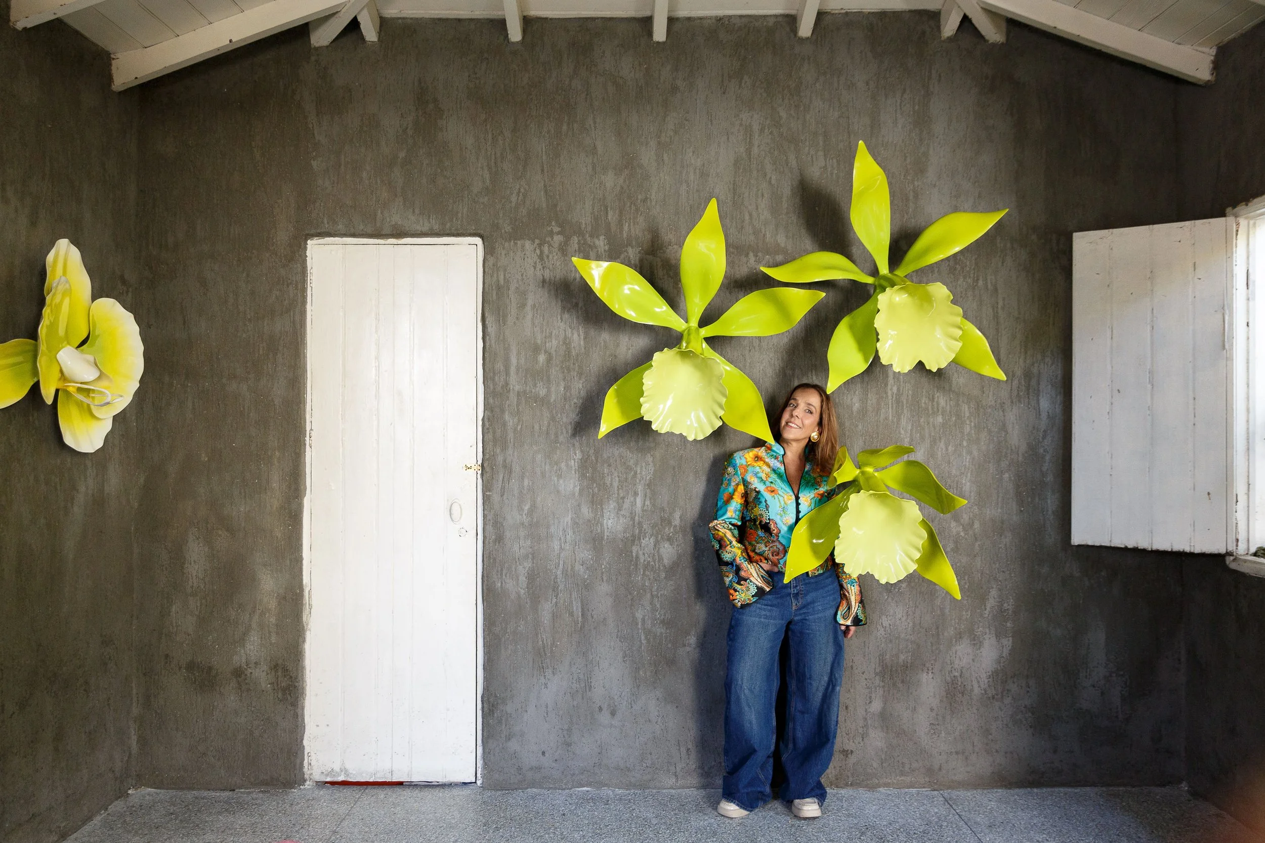 Woman in colorful patterned jacket and jeans standing against a textured gray wall background with large green plant leaves and white shutters.