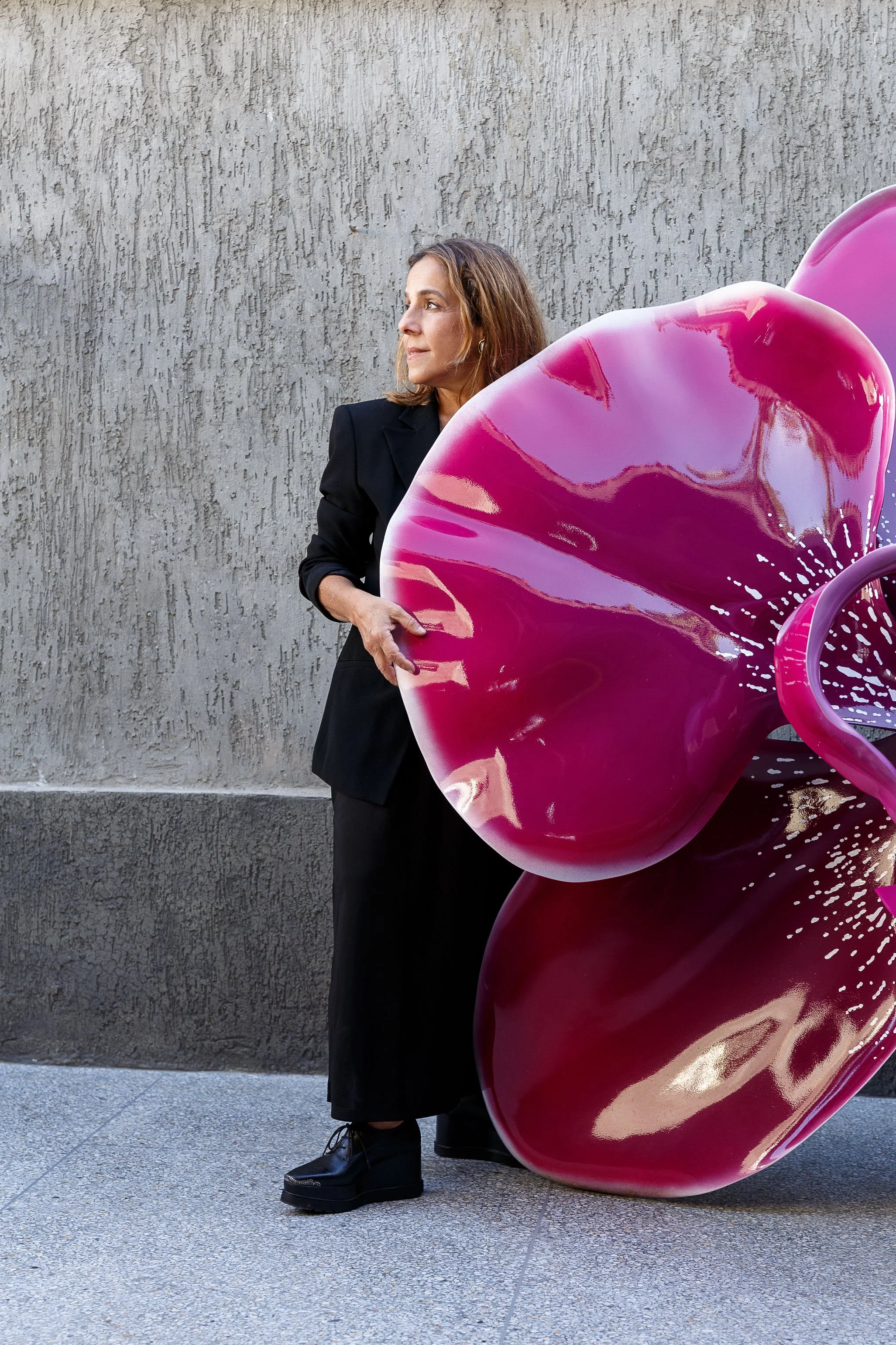Woman in black outfit standing beside a large pink flower sculpture against a textured gray wall.