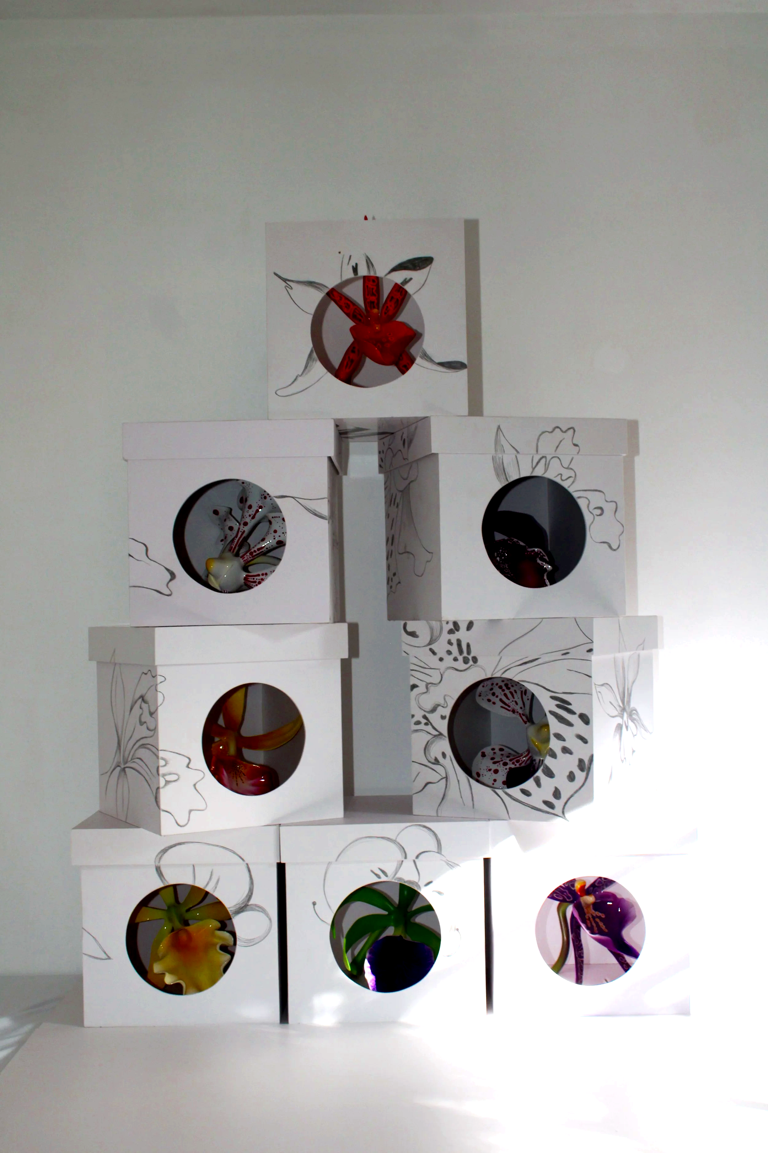 Display of several colorful glass objects inside white boxes with circular openings and black and white floral line drawings on the boxes, arranged in a pyramid, against a plain white wall.