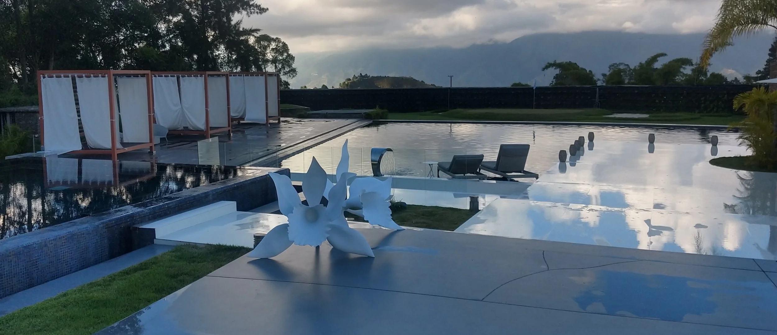 Luxury outdoor pool area with lounge chairs, cabanas with curtains, decorative white flower sculpture, and scenic mountain view at dusk.