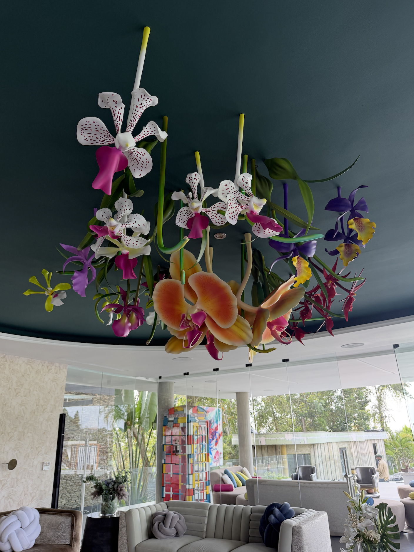 A large floral ceiling sculpture featuring orchids in pink, white, purple, and yellow hangs from a dark ceiling in a modern room with large glass windows and furniture.