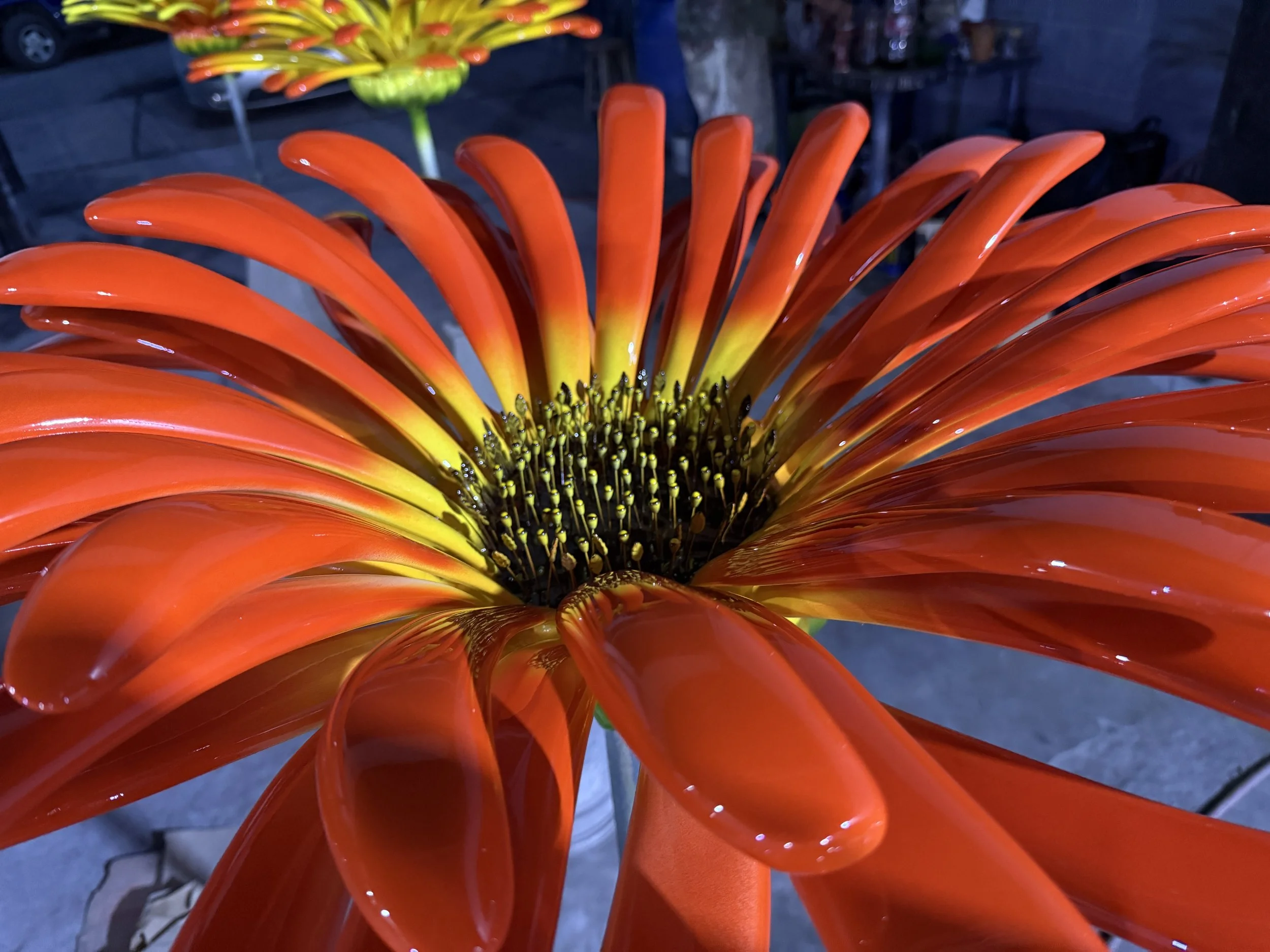 Close-up of a vibrant orange and yellow flower sculpture with long, curved, glossy petals and a dark center. The background shows an outdoor setting with a parking lot and other flower sculptures.