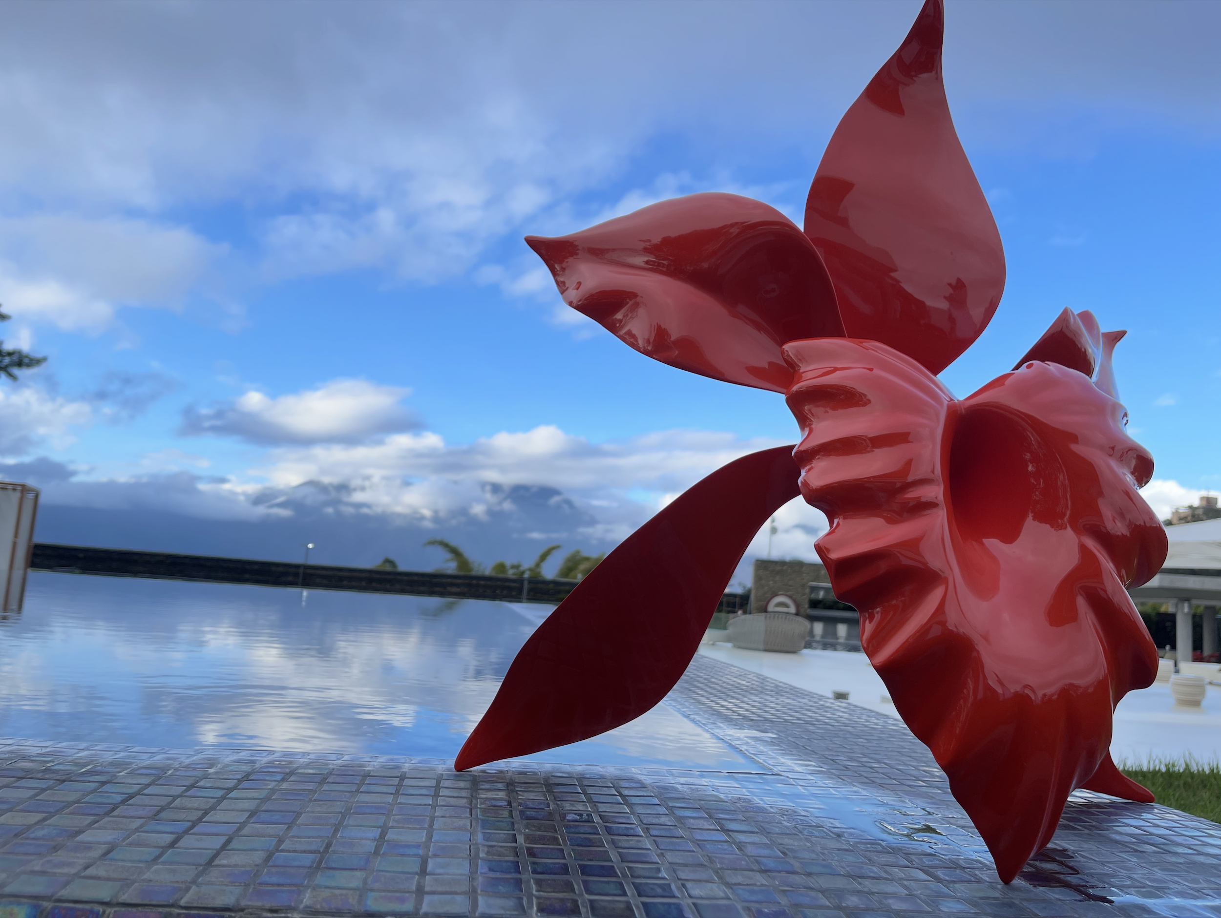Red abstract sculpture with curved shapes outdoors against a blue sky with clouds, near a pool or water feature.