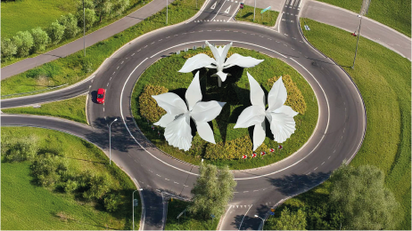 A roundabout with large white flower sculptures in the center, surrounded by green grass and trees.
