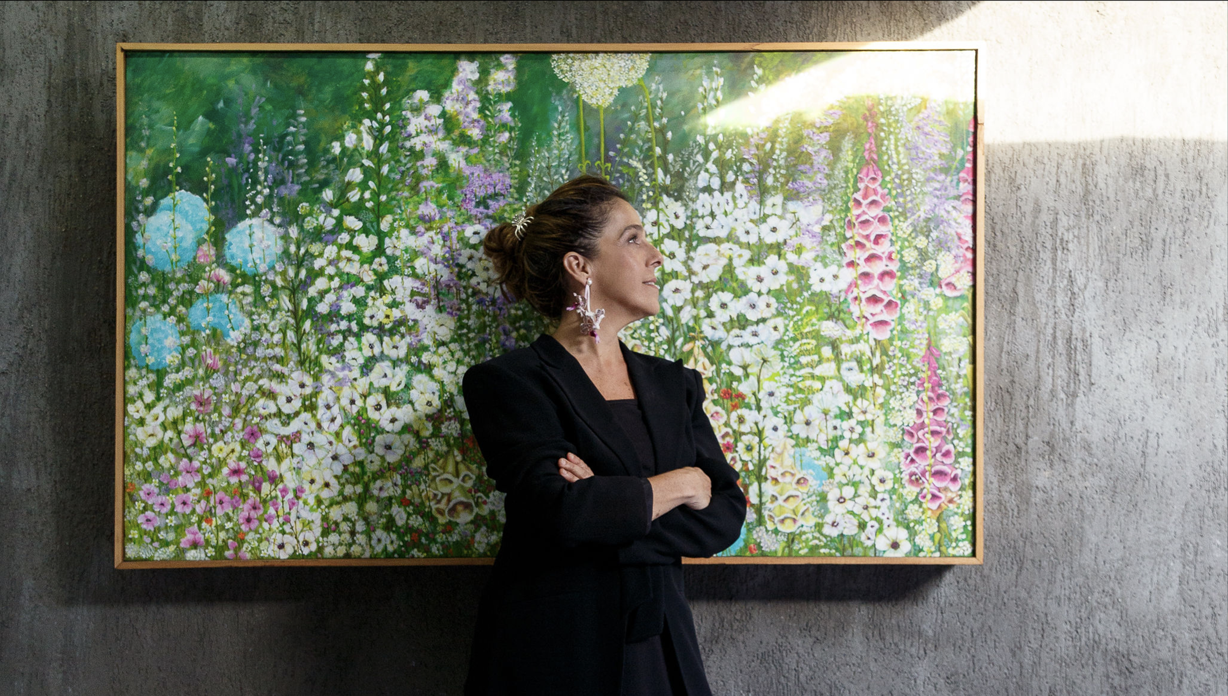 A woman stands with arms crossed in front of a colorful floral painting on a gray concrete wall. Sunlight shines on the painting from the right side.