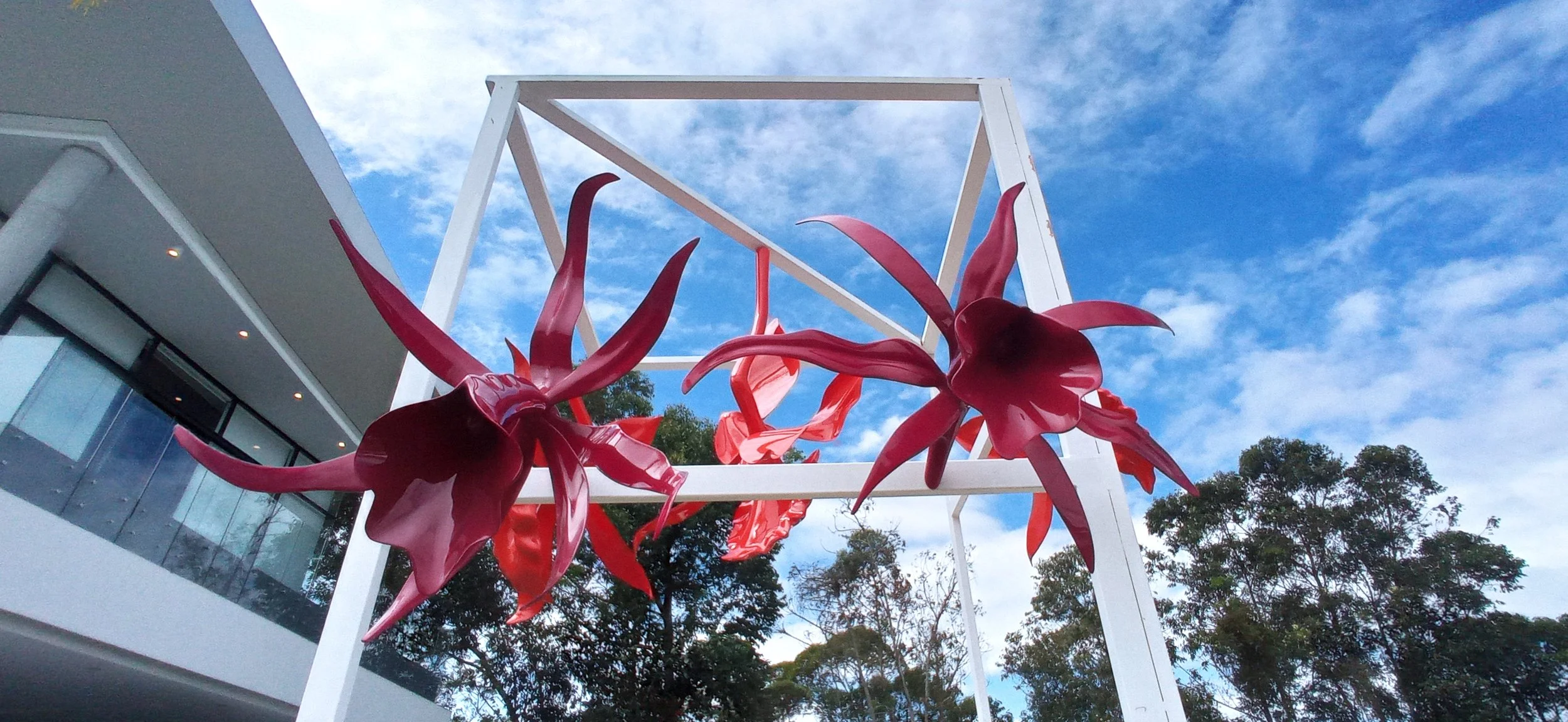 Red glass floral sculpture mounted on a white geometric frame outdoors, with a building and trees in the background under a partly cloudy sky.