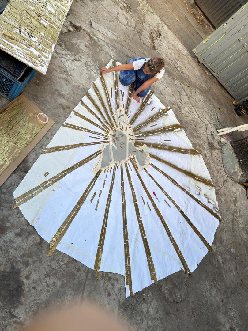 A woman sitting on the ground assembling a large, circular mosaic or decoration made of strips of material, possibly metal or reflective tape, arranged in a radial pattern. The mosaic features a religious or religious-like design with a central figure. She is working outdoors on a concrete surface with various materials and tools around her.