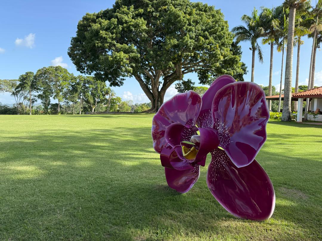 A large purple orchid sculpture on a grassy lawn with trees, palm trees, a small building with a red roof, and blue sky in the background.