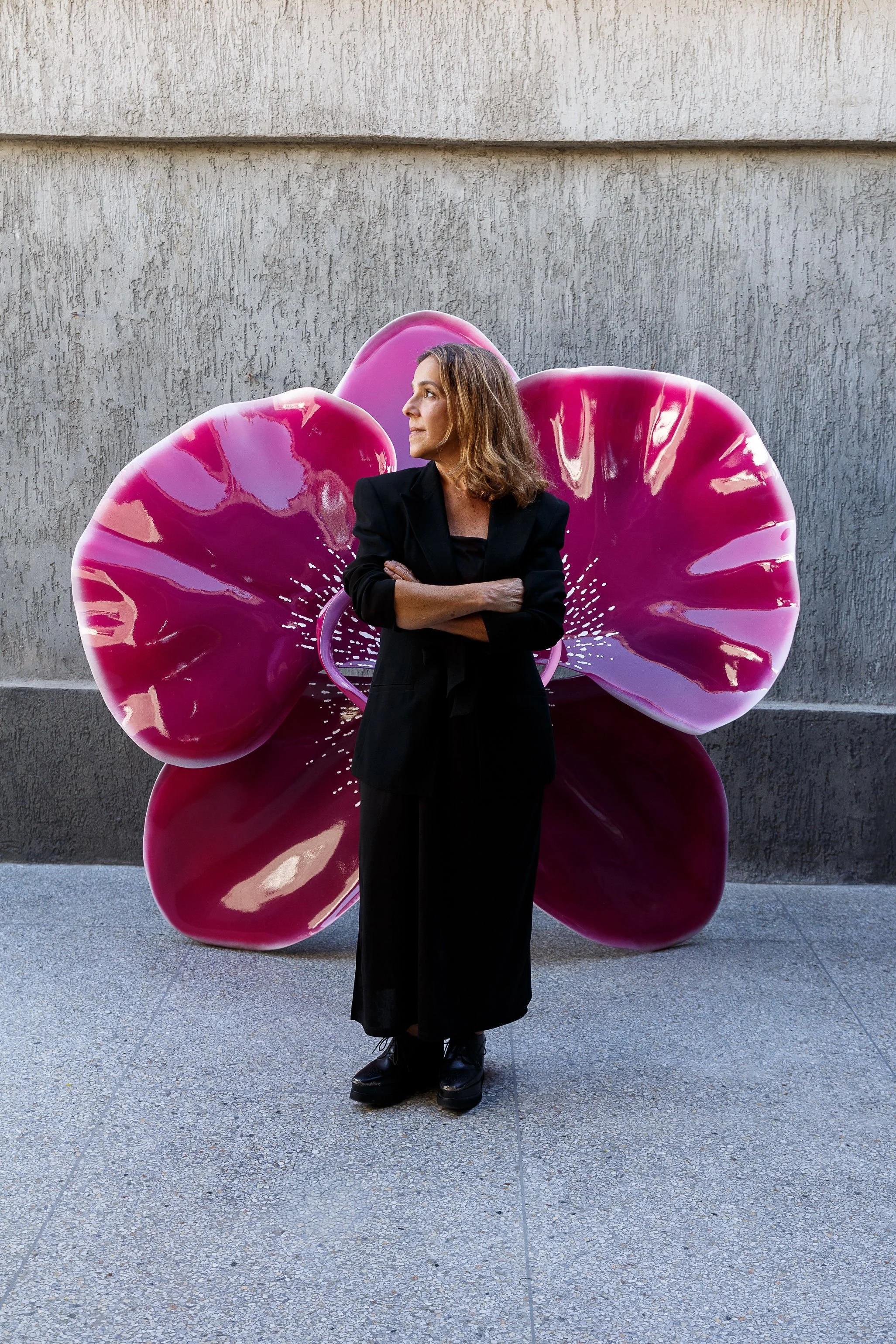 A woman with shoulder-length brown hair stands with her arms crossed in front of a large, pink, flower-shaped sculpture attached to a textured concrete wall.