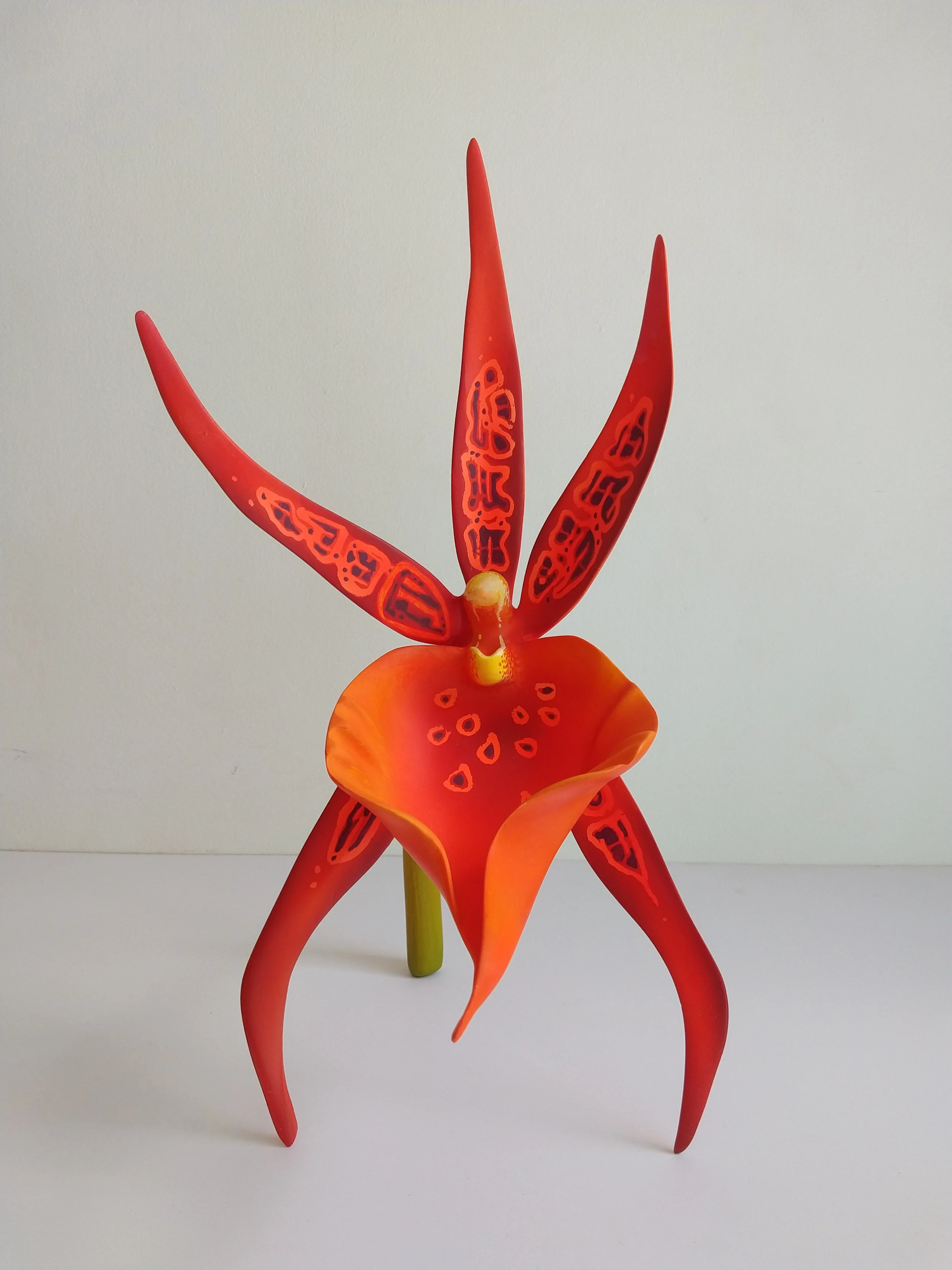 Artificial red and orange flower sculpture with long, pointed petals and a green stem, set against a plain white background.