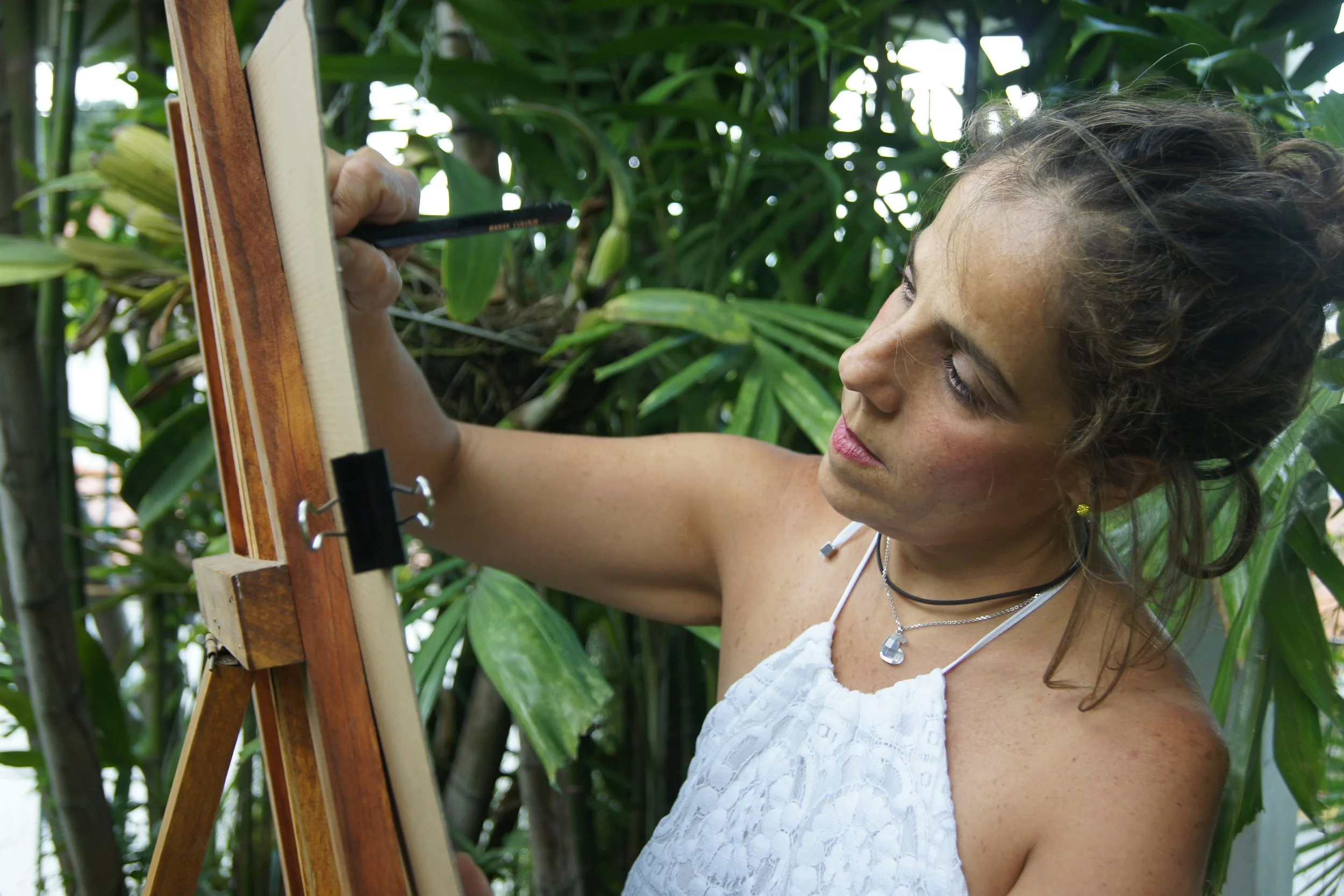 A woman with curly hair and wearing a white sleeveless top is painting on an easel outdoors surrounded by green plants.