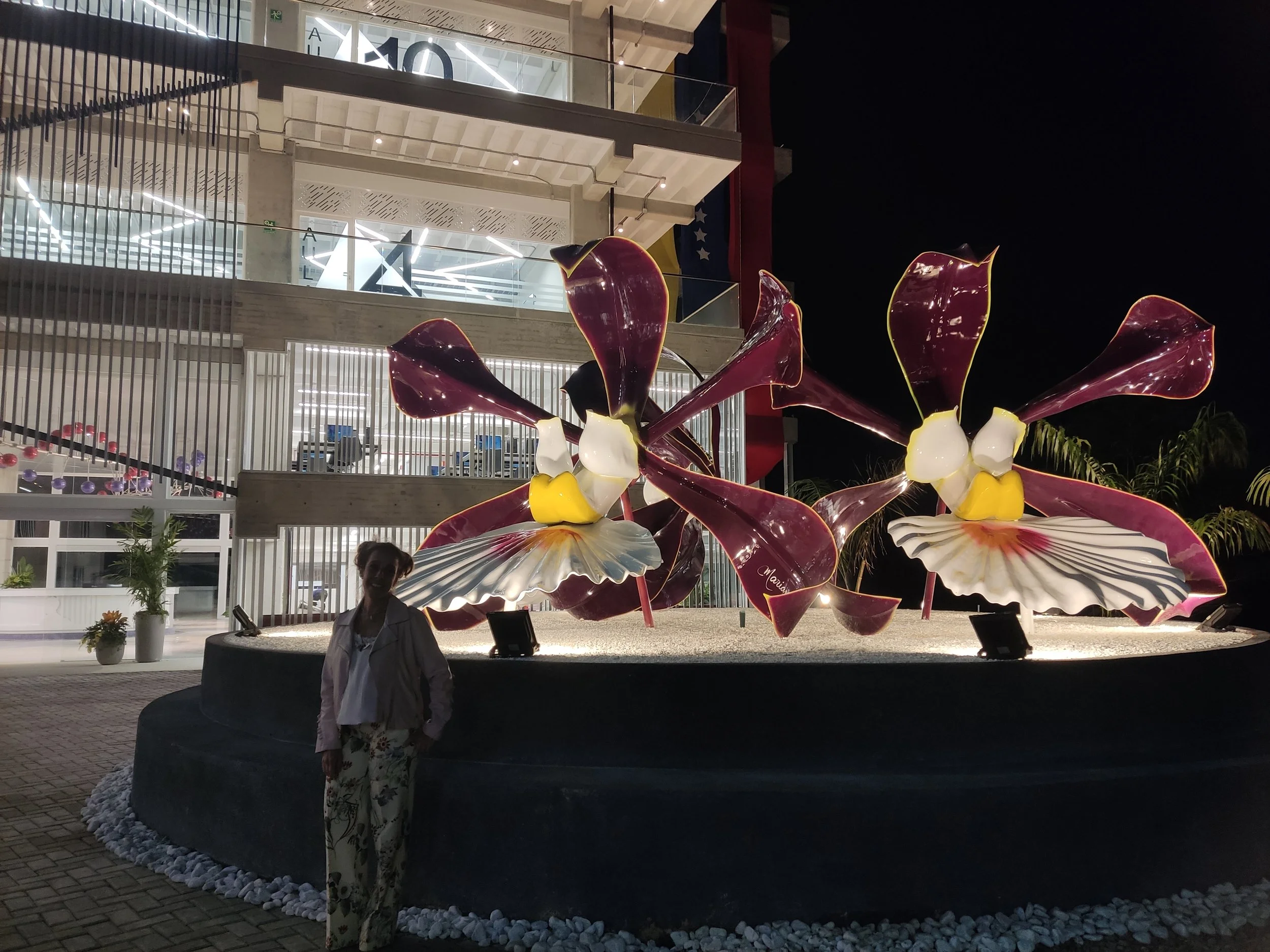 Night view of a modern building with glass balconies and illuminated interior, decorated with large glass sculptures resembling flowers with purple, yellow, and white petals, in an outdoor area with a woman standing in front.