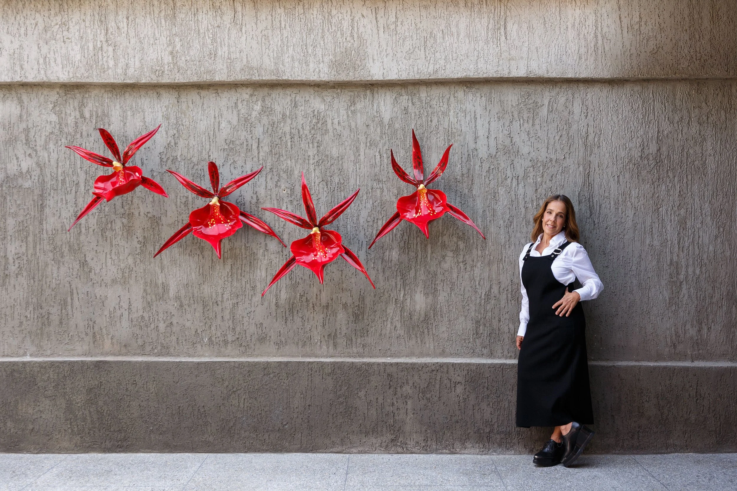 A woman with light brown hair wearing a black pinafore dress over a white shirt, standing against a beige textured wall. To her left, there are three large red artificial flowers mounted on the wall.