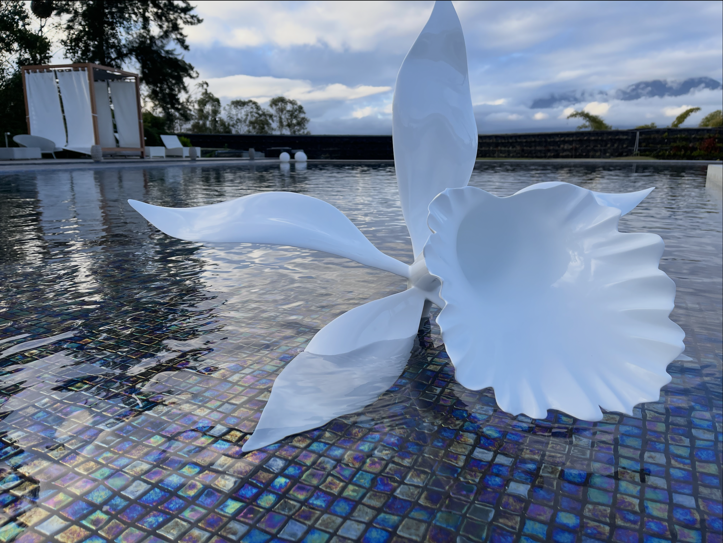 Close-up view of a white decorative sculpture resembling a flower or abstract shape floating on a pool with iridescent mosaic tiles, with outdoor lounge chairs and a wooden cabana in the background under a partly cloudy sky.
