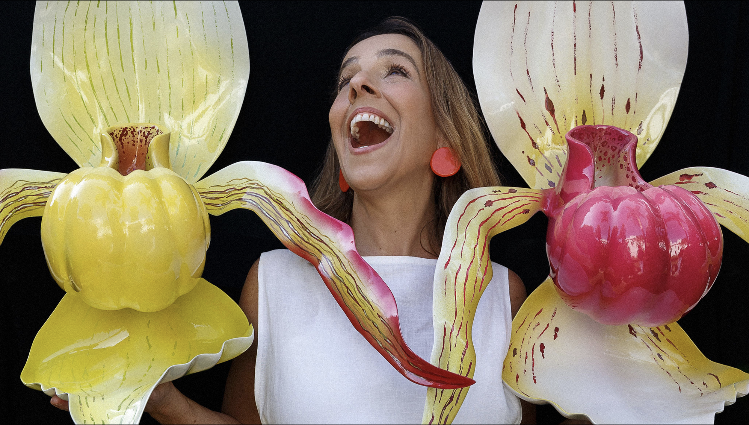 A woman with long brown hair, wearing a white dress and orange earrings, is smiling widely and looking upward. She is surrounded by large, colorful, decorative orchid flower sculptures in yellow and pink with green and red accents.