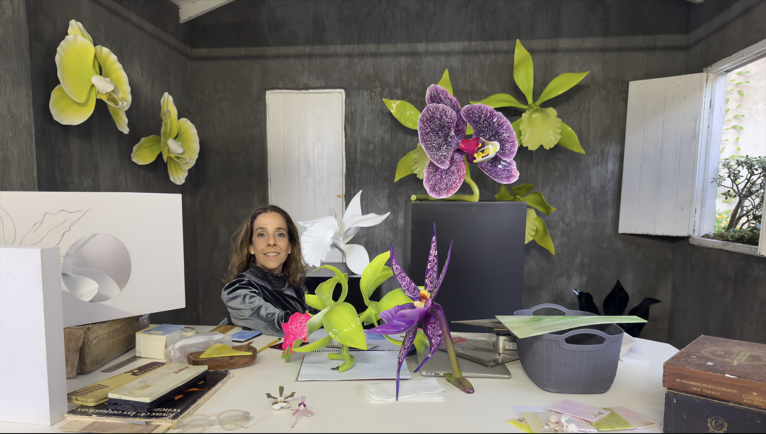 Woman sitting at a desk surrounded by colorful artificial flower sculptures, with wall art and a window in the background.
