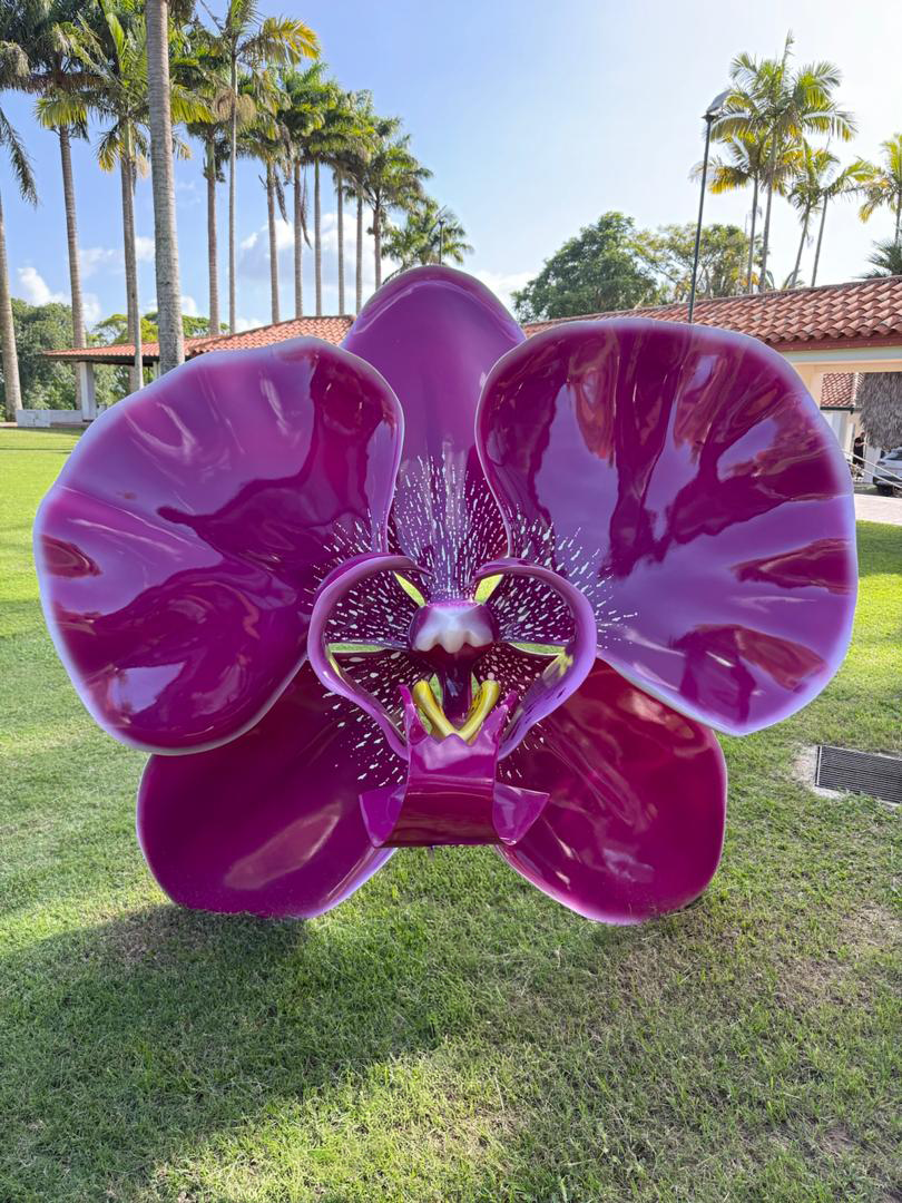 Large, purple, orchid-shaped sculpture with reflective petals and detailed inner parts, placed on a grassy area outdoors with palm trees and a building in the background.