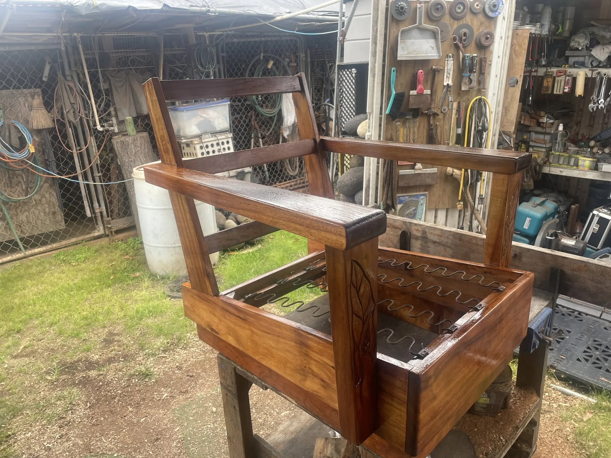 Unfinished wooden chair frame sitting outdoors on a workbench with a cluttered workshop background.