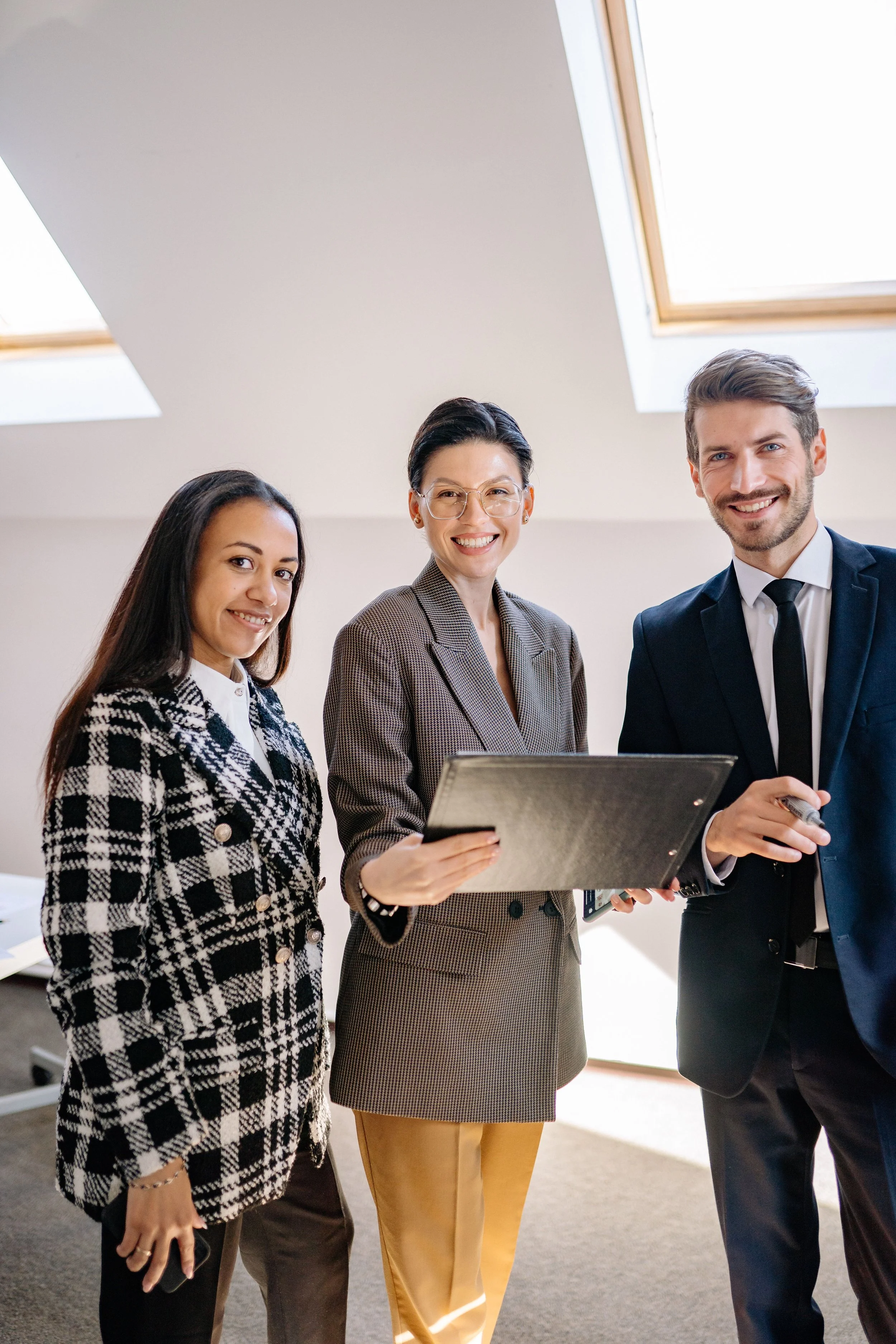 Three professionally dressed individuals smiling in an office with skylight windows.