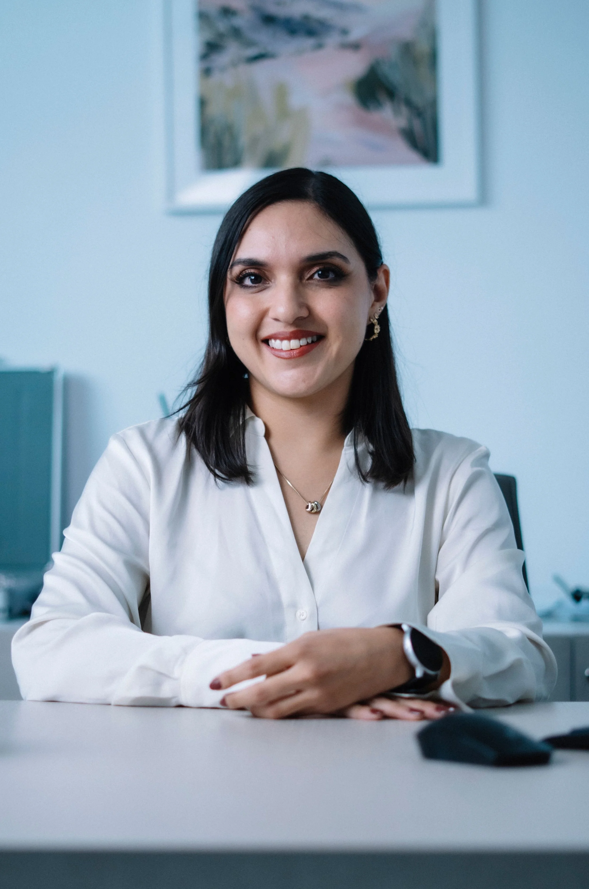 A smiling woman with dark hair and earrings, dressed in a white blouse, sitting at a table with her hands resting on it, in an office setting.