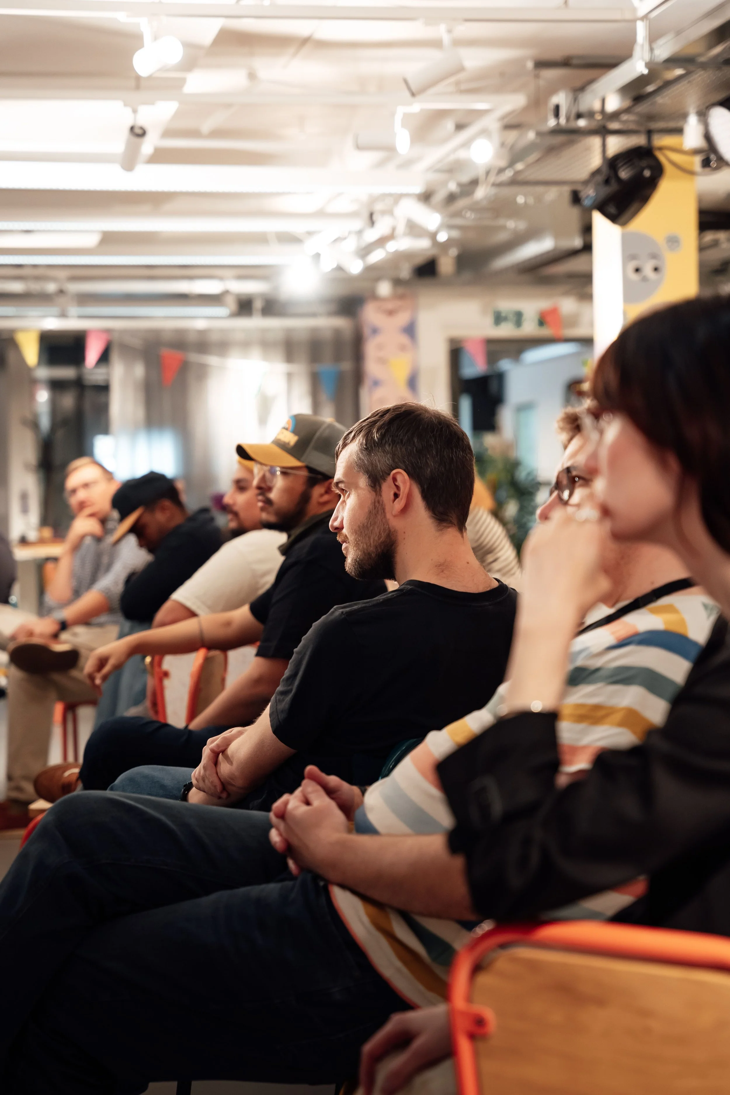 A group of people sitting indoors, attentively listening or watching something. The setting appears to be a casual or community event, with a diverse group of individuals seated in a row.