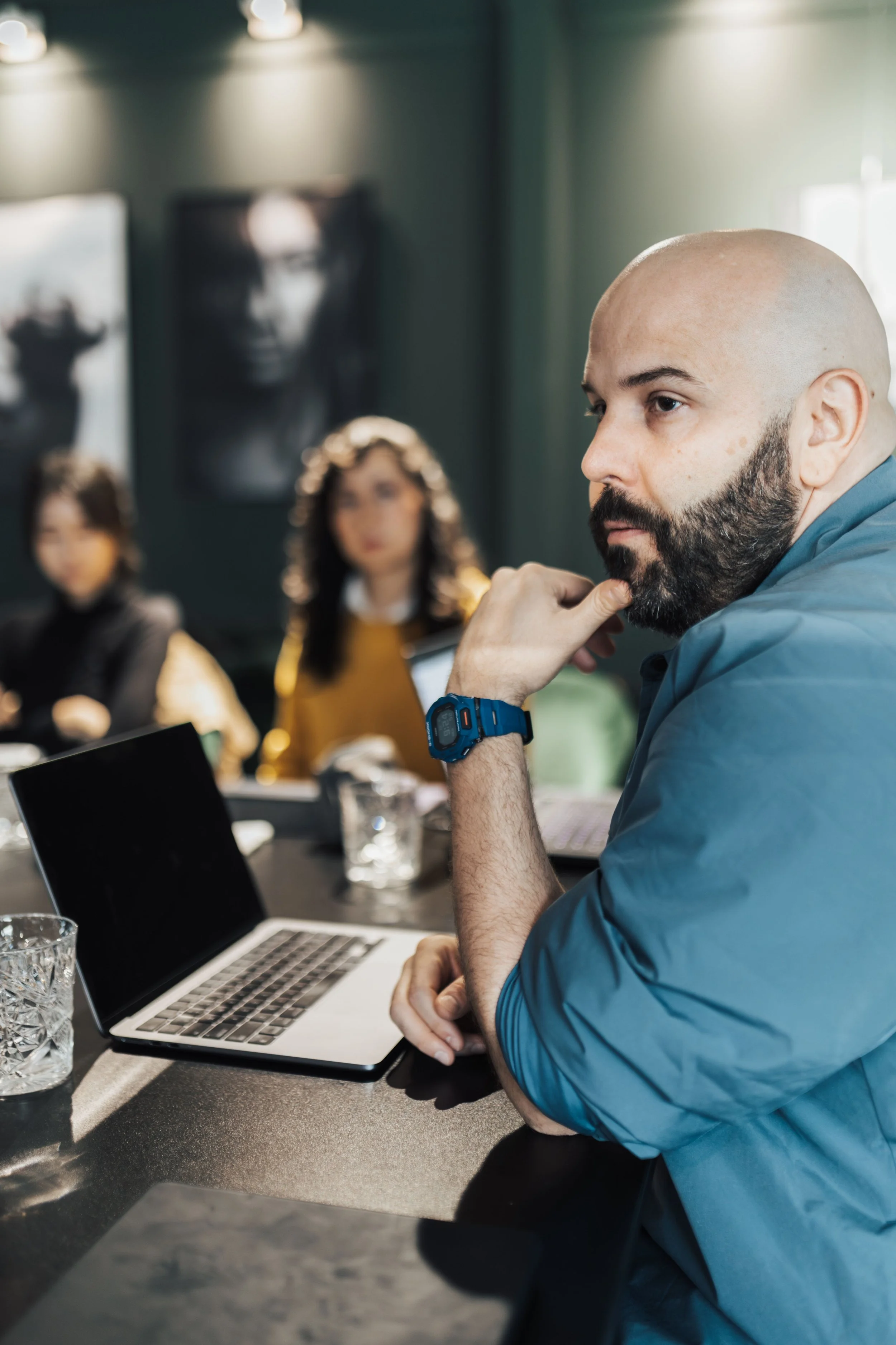 A man with a shaved head and beard sitting at a table with an open laptop, drinking water, with glasses and a blurred background of other people in a meeting room.