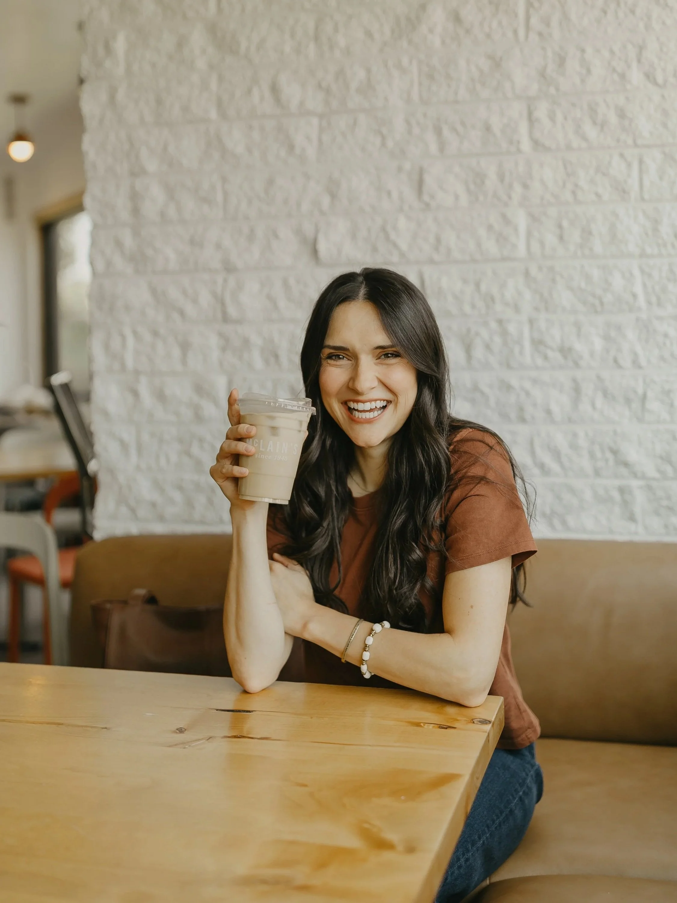Young woman with long dark hair, smiling and holding a large iced coffee, sitting at a wooden table in a cafe with a white brick wall background.