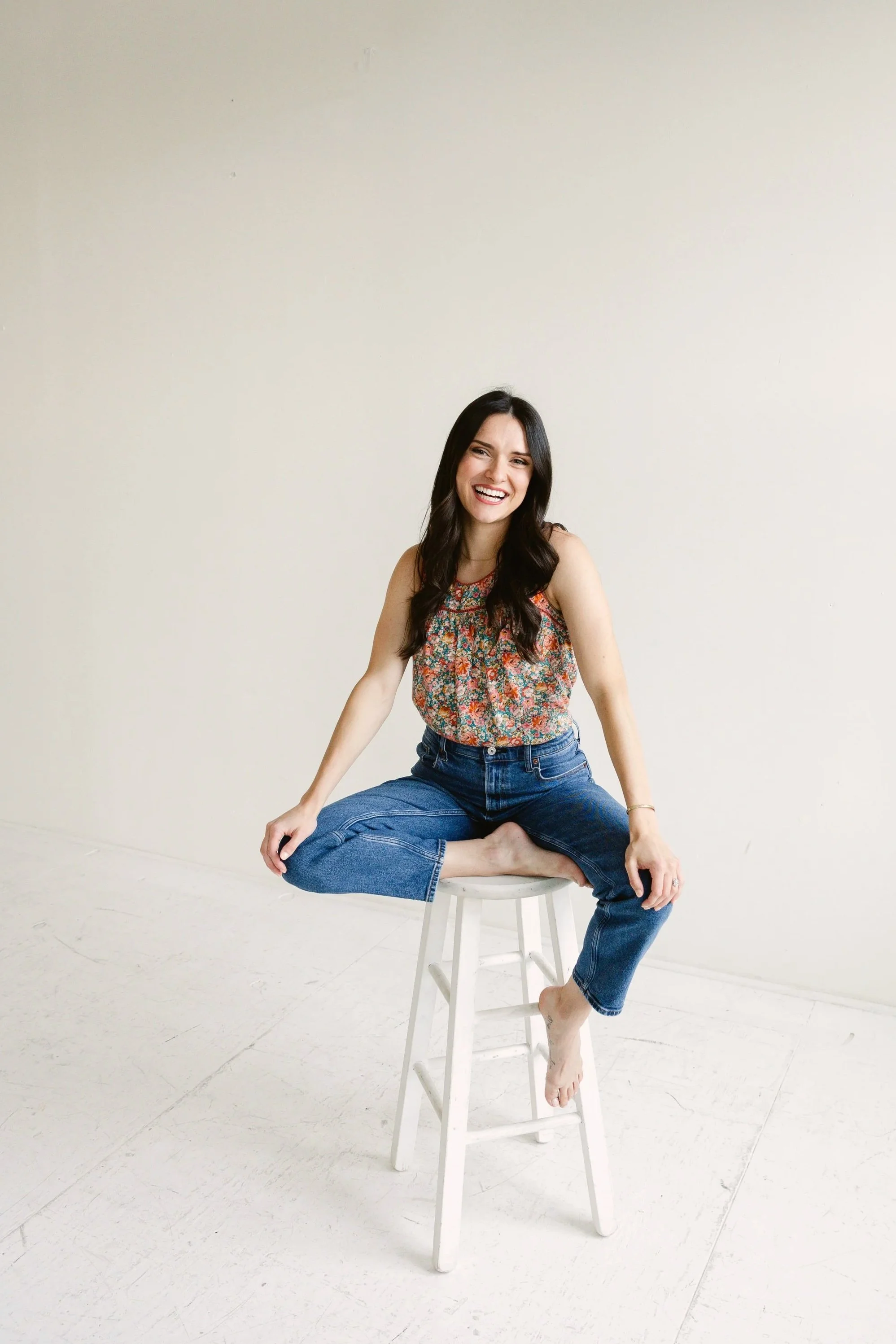 A woman with long dark hair sitting on a stool with legs crossed, smiling, wearing a floral sleeveless top and blue jeans, barefoot, against a plain cream-colored background.