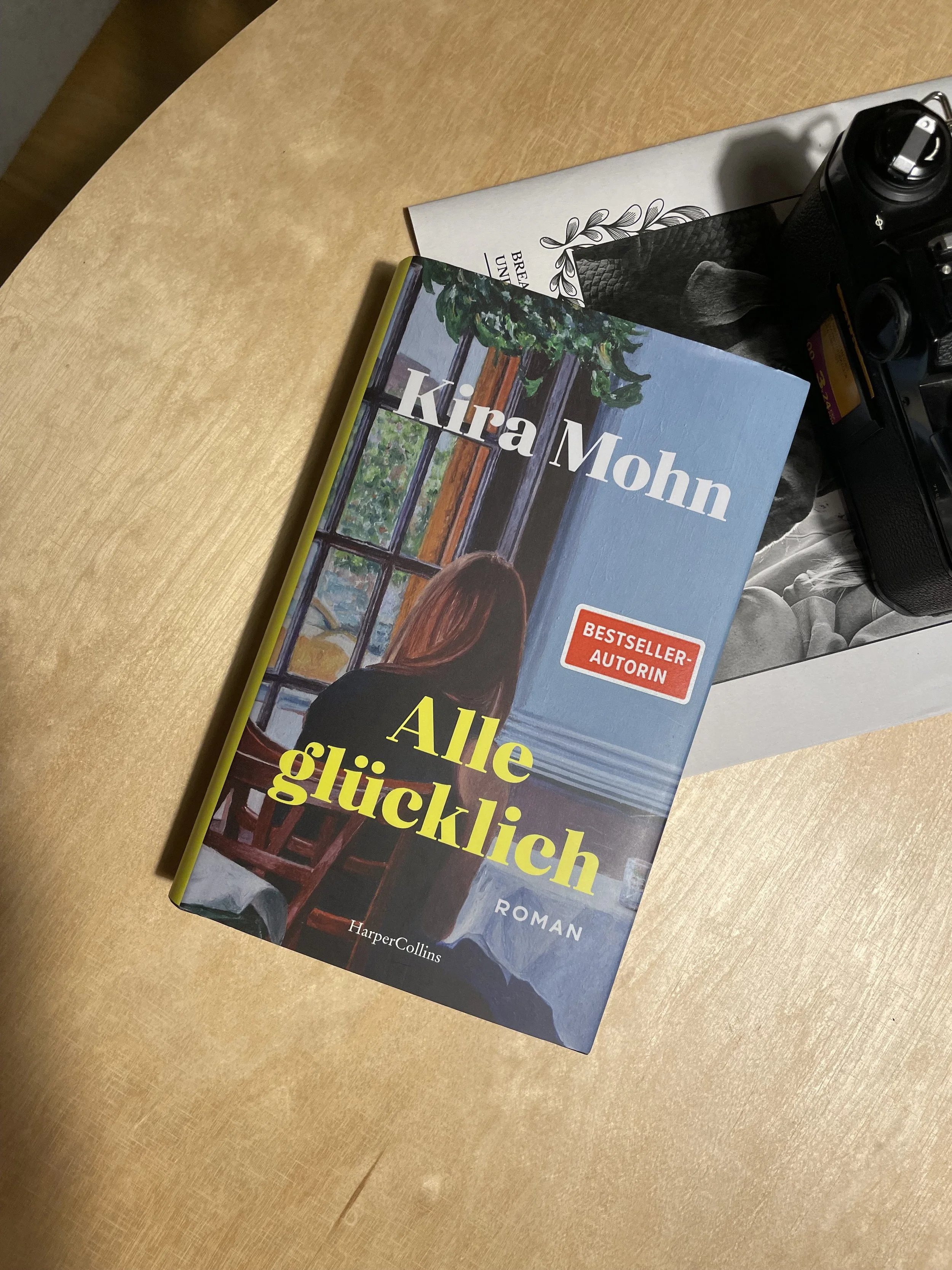 Auf einem Holztisch liegen der Roman "alle glücklich" von Kira Mohn, eine schwarze Analog Nikon Kamera und eine Zeitung.