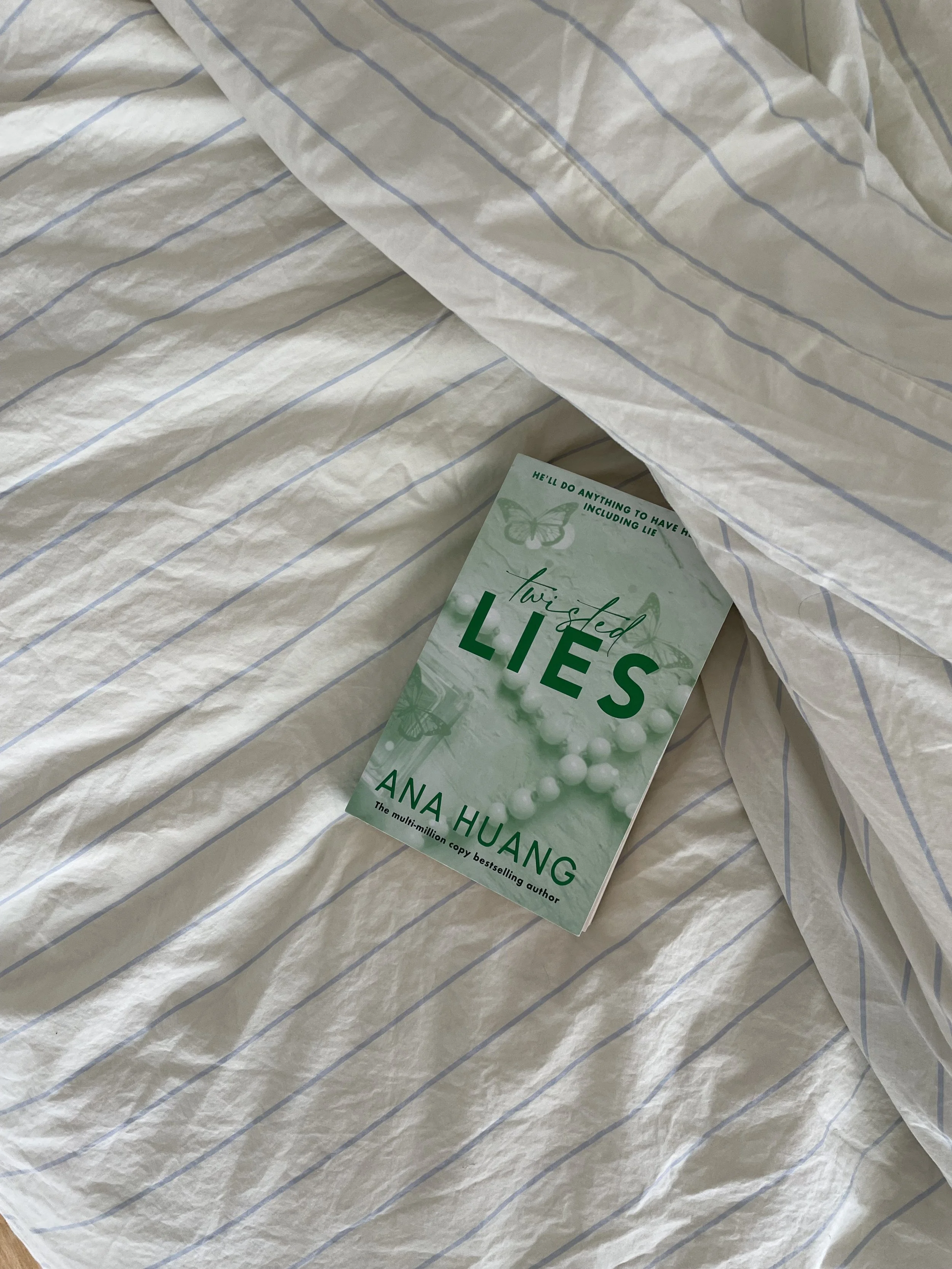Das Buch „Twisted Lies“ von Anna Huang liegt auf einer weiß-grau gestreiften Bettdecke.
