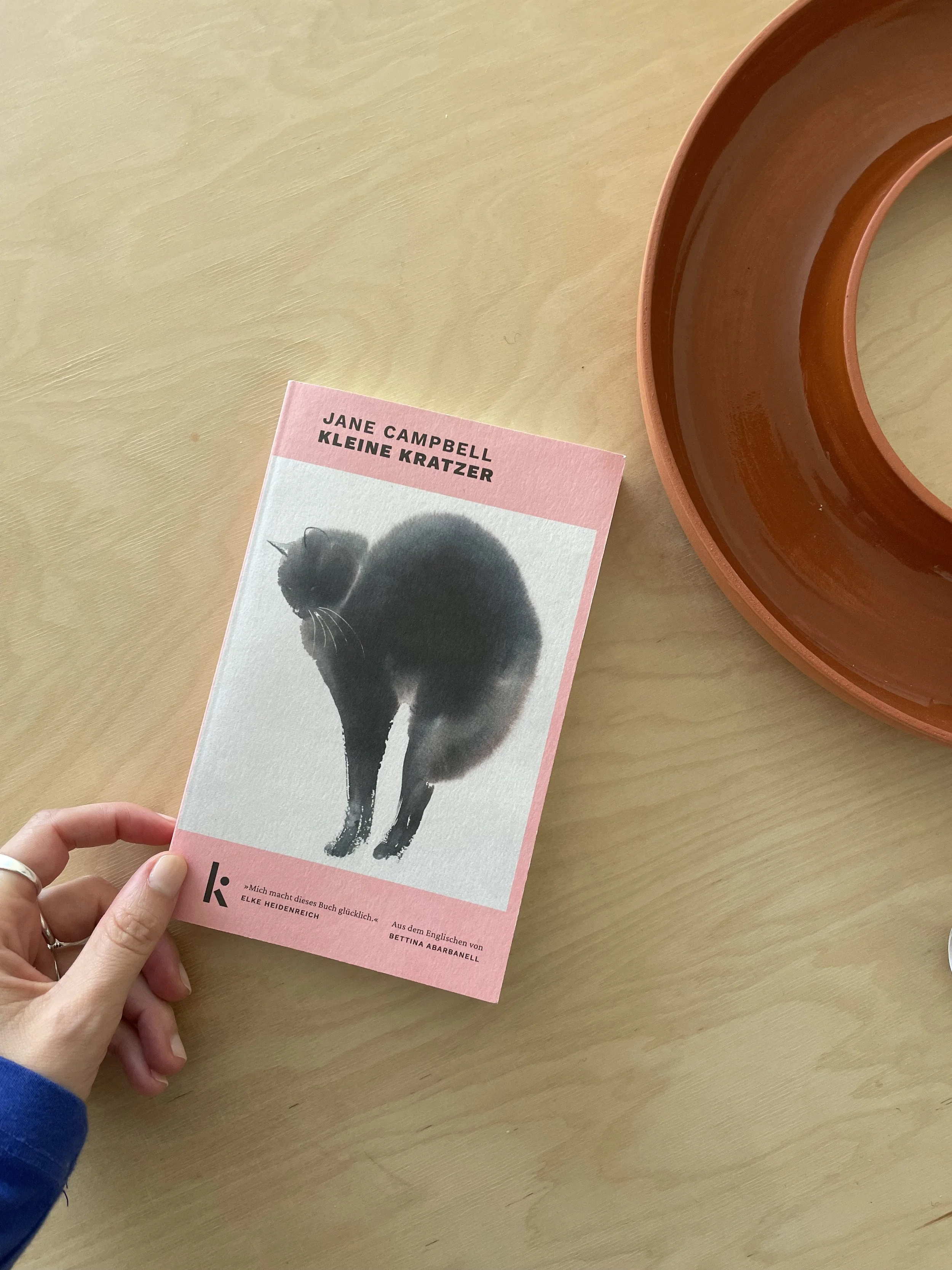 Das Buch „Kleine Kratzer“ von Jane Campbell mit einer schwarzen Katze auf dem Umschlag liegt neben einer orangefarbenen Keramikschale auf einem Holztisch.
