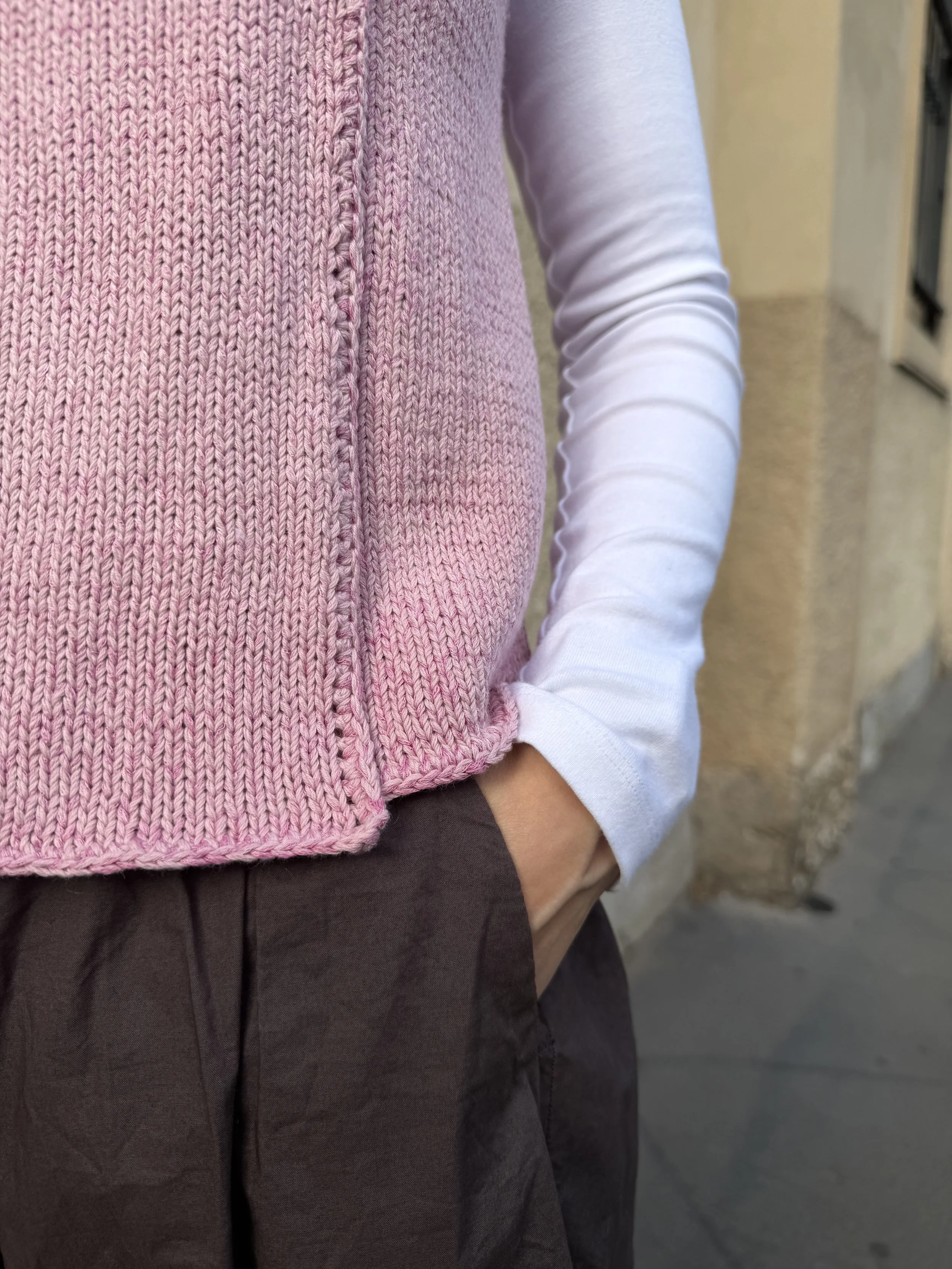 Nahaufnahme einer handgestrickten rosa Weste mit asymmetrischem Schnitt und gerippten Bündchen über einem weißen Langarmshirt.