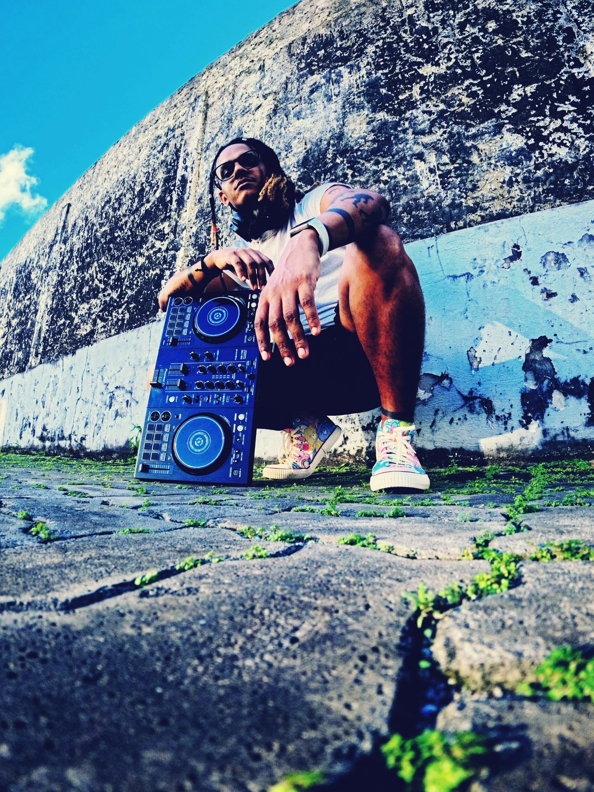 A person crouching on a cobblestone street with small patches of grass, holding DJ equipment, in front of a weathered white wall with peeling paint, under a clear blue sky.