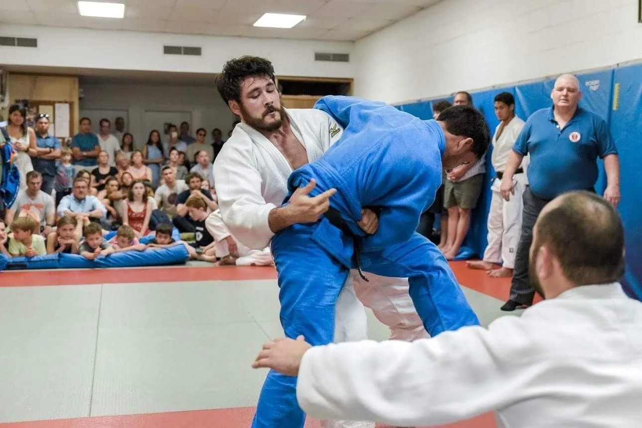 Two judo competitors in white and blue gi engaged in a match on a matted floor, with an audience watching in the background and a referee observing.