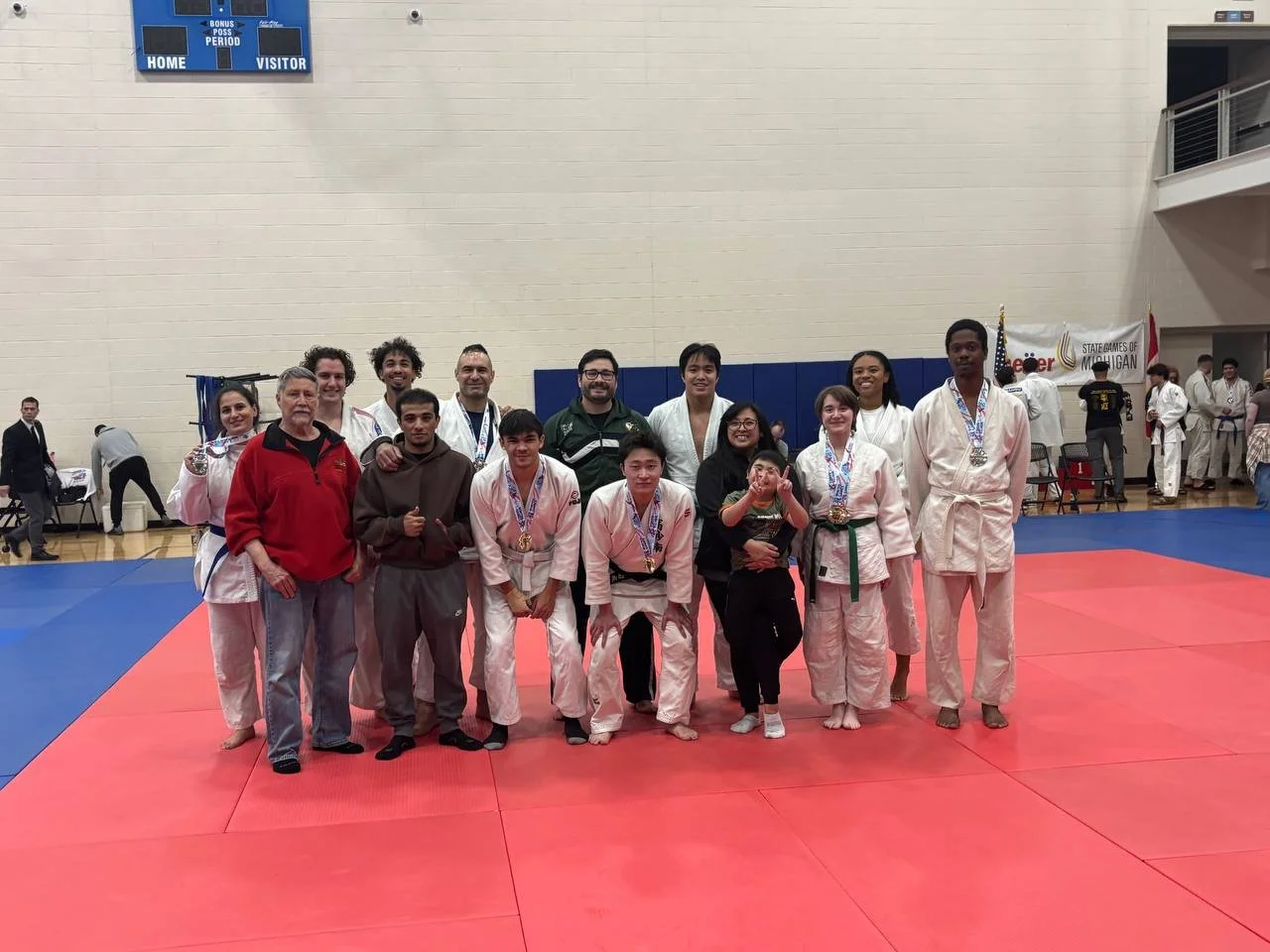 Group of judo athletes and coaches posing together on a red and blue tatami mat in a sports hall, wearing medals and judo uniforms.