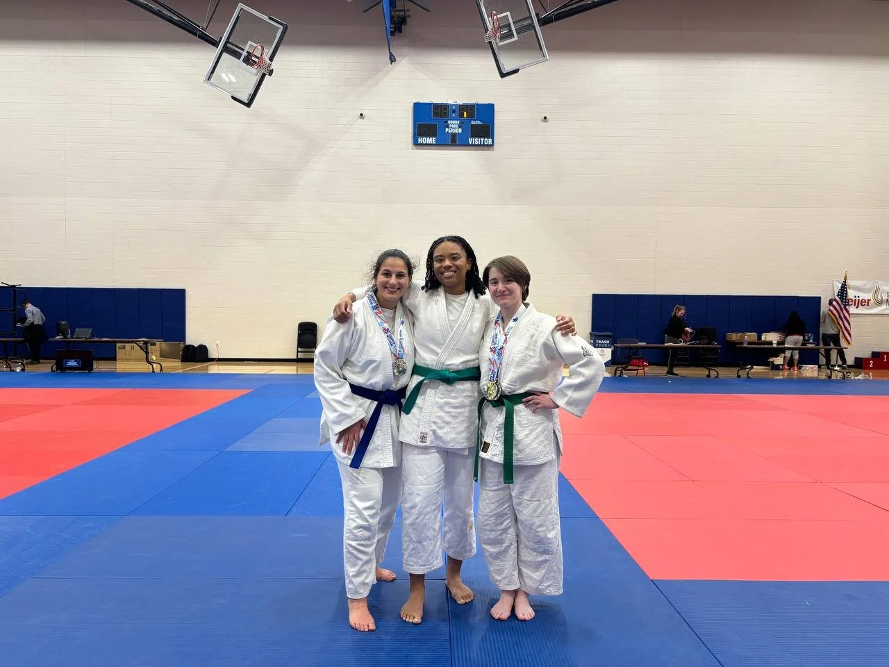 Three female martial artists in judo gis with medals around their necks, standing on a red and blue tatami mat in a gymnasium after competition.