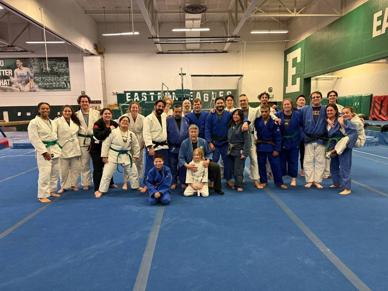Group of judo practitioners and instructors, including children and adults, posing together on a blue mat inside a gymnasium after training, wearing traditional judo uniforms.