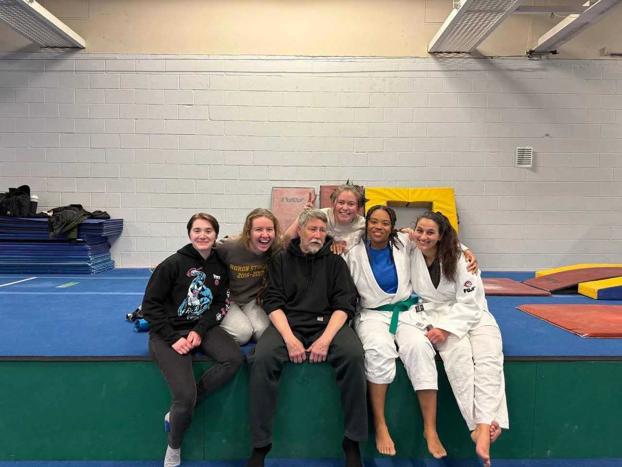 Group of five people sitting on a green mat in a gym, some wearing martial arts uniforms, smiling and posing for a photo.