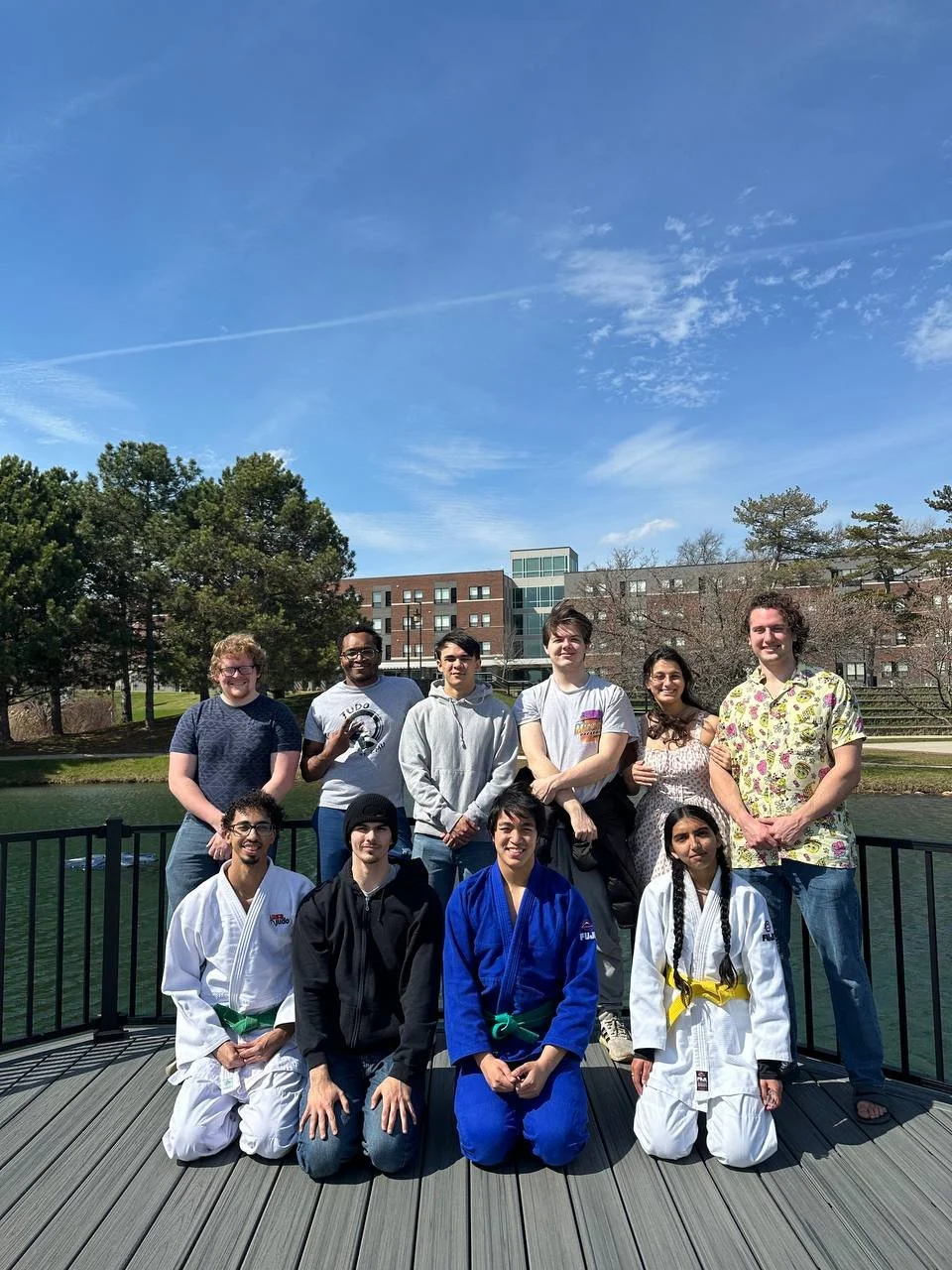 Group of people outdoors on a wooden platform near a body of water, with some people in martial arts uniforms with colored belts, others in casual clothing, with trees and buildings in the background under a partly cloudy blue sky.