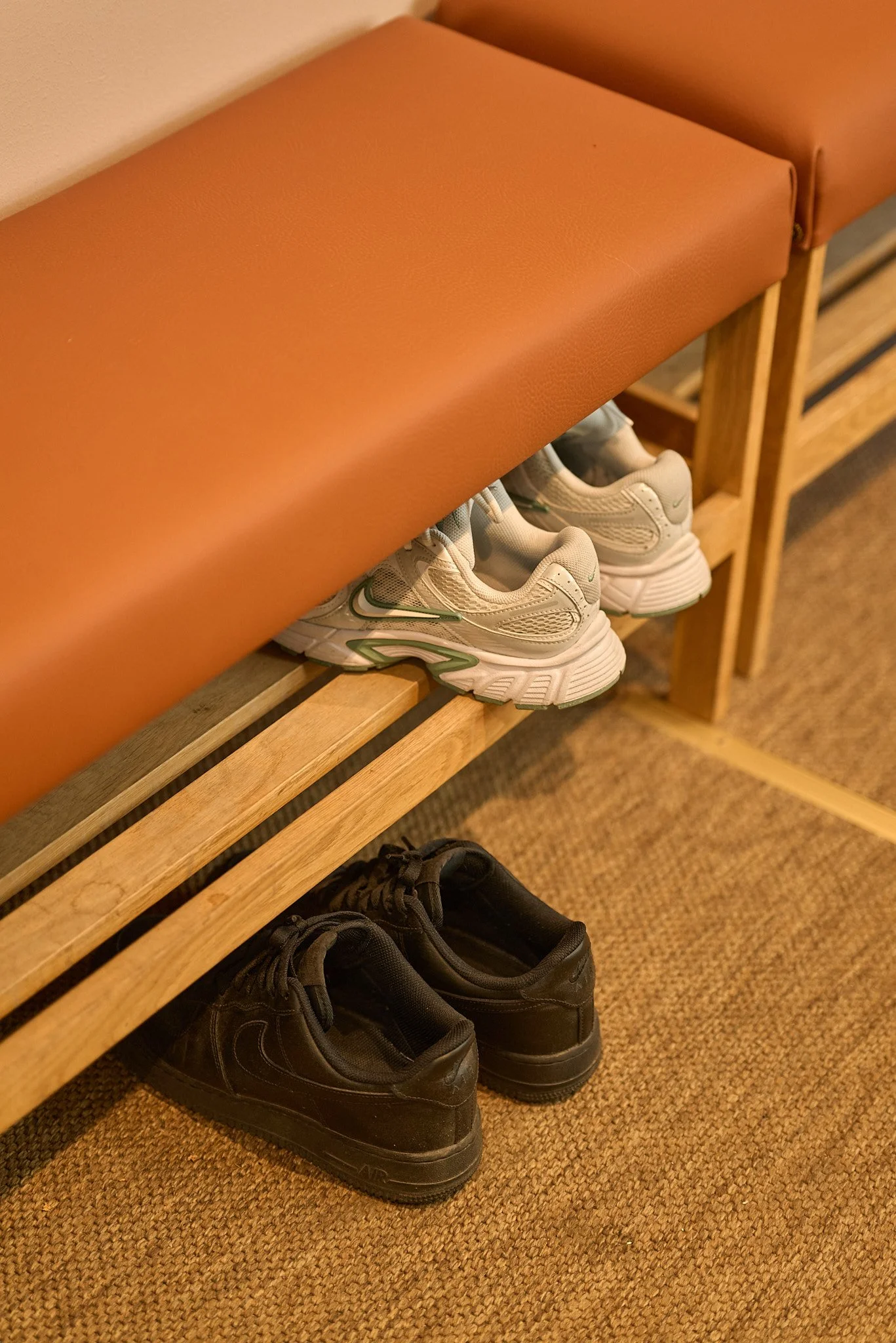 Pair of white athletic shoes and pair of black athletic shoes on a wooden bench with a brown cushioned top, in a room with a brown carpeted floor.