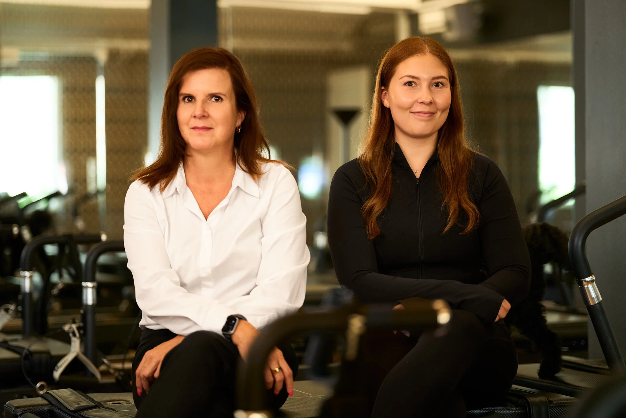 Two women sitting on chairs in what appears to be an airport or train station lobby, with luggage around them. The woman on the left has brown hair, is wearing a white shirt, and has a smartwatch on her left wrist. The woman on the right has long red hair, is wearing a black zip-up jacket, and has her arms crossed. Both women are smiling and looking at the camera.