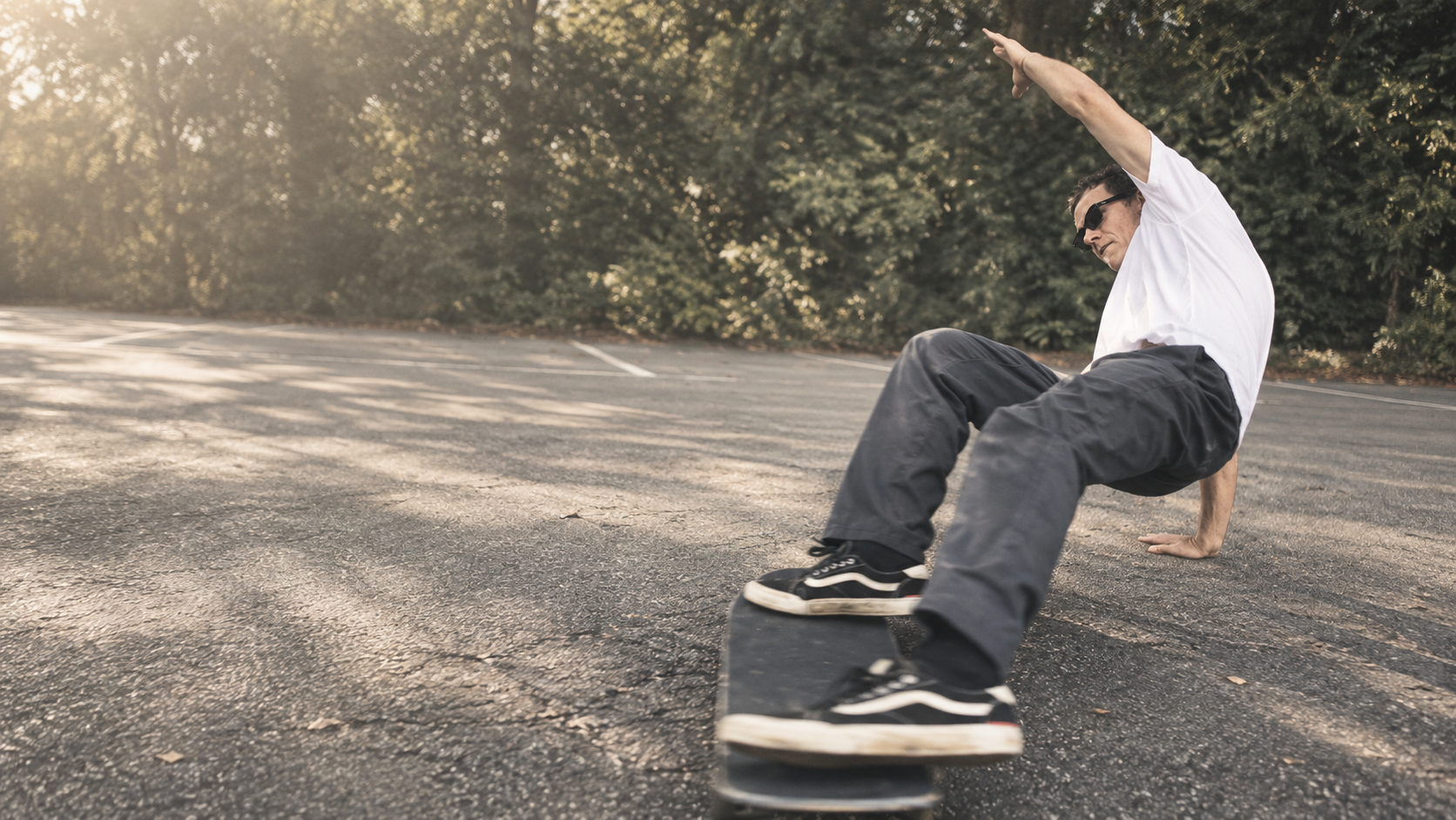 A man skateboarding on an empty asphalt road with trees and sunlight in the background.