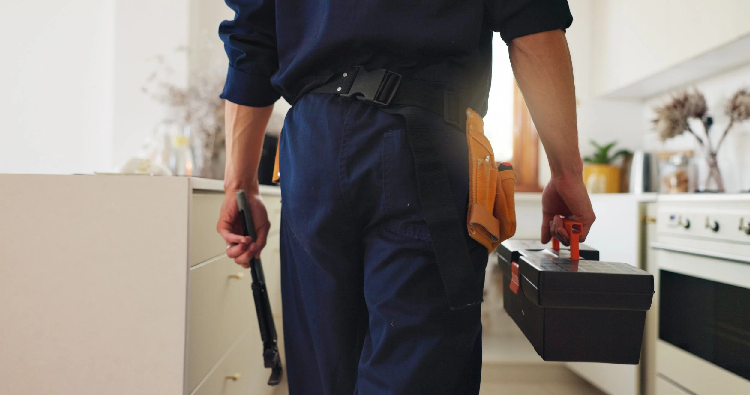 A person wearing a black shirt and navy pants with a tool belt is holding a cordless drill in one hand and a small black toolbox with an orange handle in the other, standing in a kitchen.