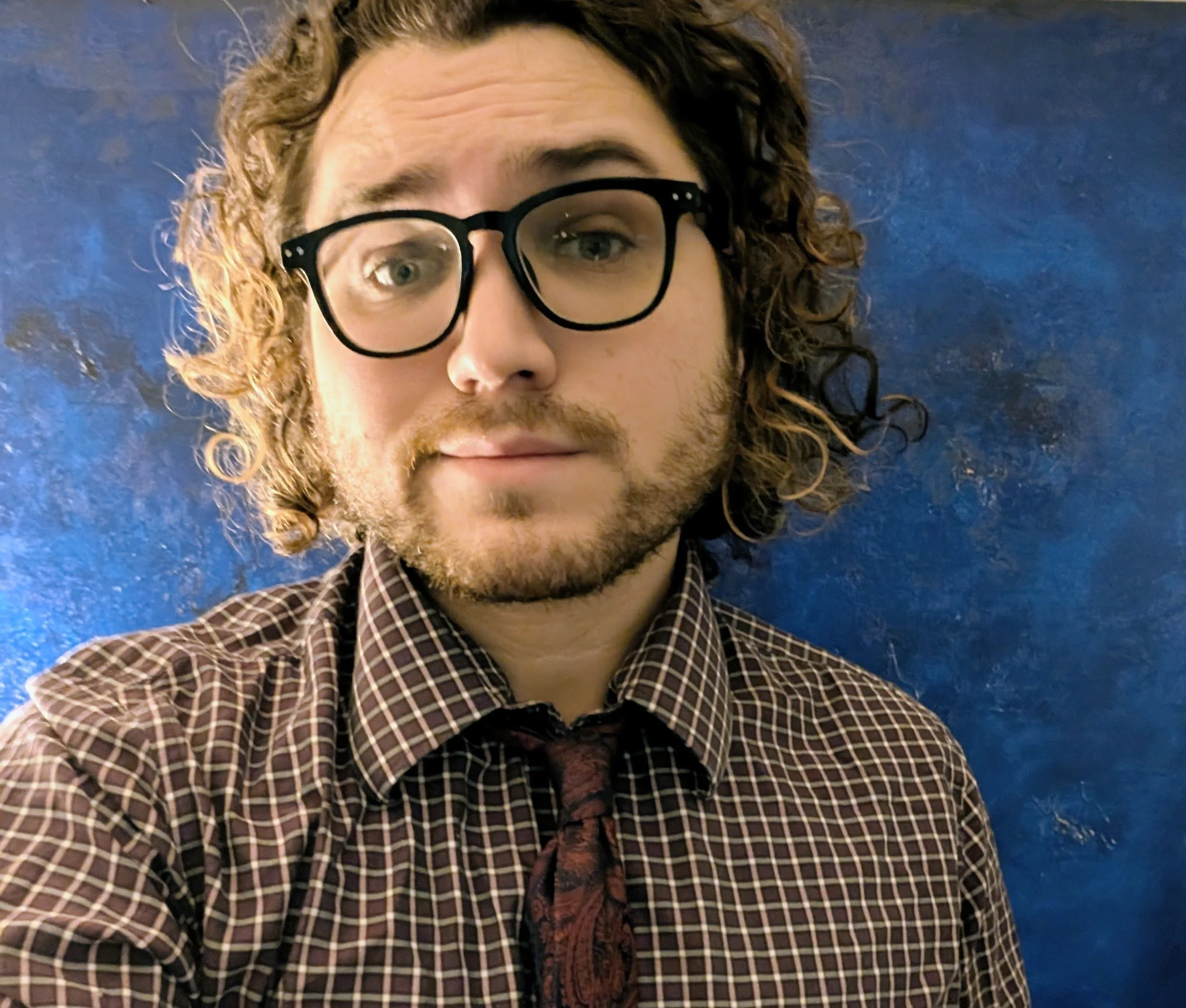 Close-up of a man with curly hair wearing large black glasses, a checkered shirt, and a dark tie, against a blue background.