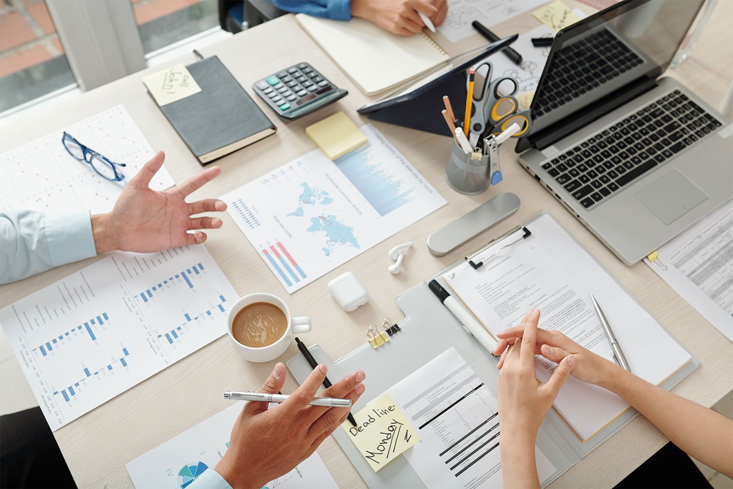 An overhead view of a cluttered office desk with two people working, one with a pen and the other gesturing, surrounded by documents, graphs, a laptop, glasses, calculator, sticky notes, a coffee cup, and office supplies.