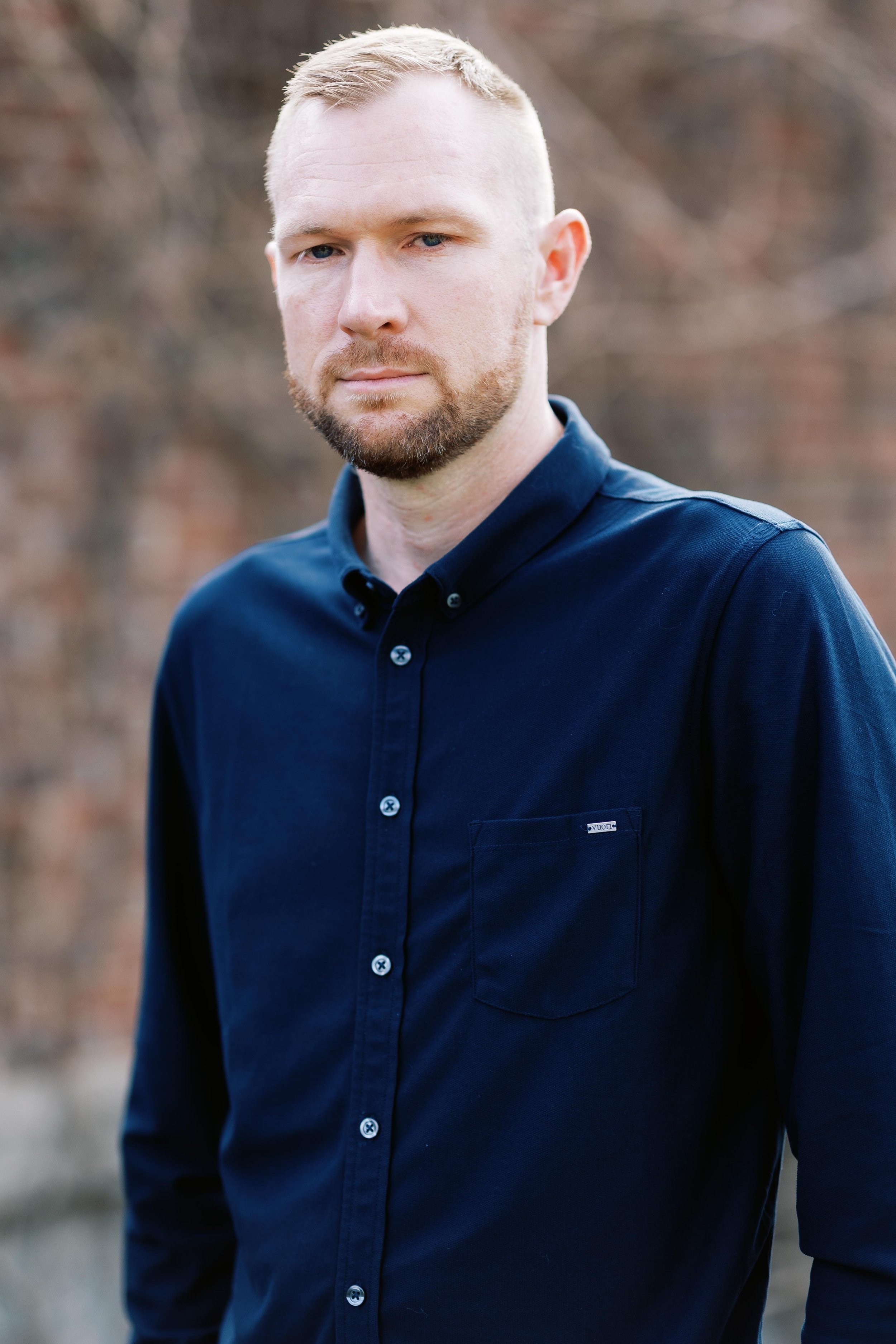 A man with short blonde hair and a beard wearing a navy blue button-up shirt, standing outdoors with a blurred natural background.